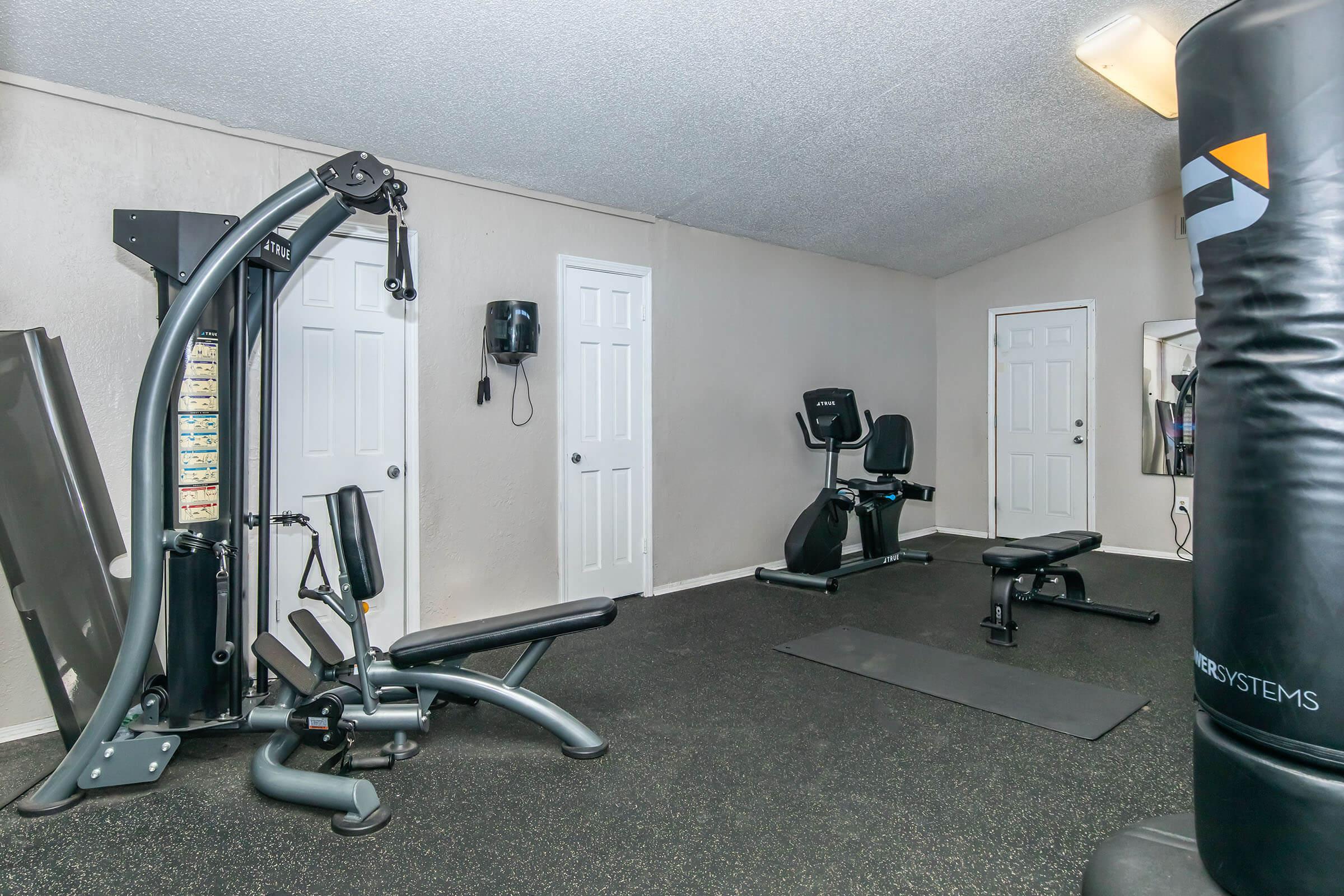 A small home gym featuring various exercise equipment, including a multi-station weight machine, a bench, a stationary bike, and a punching bag. The room has light-colored walls and black rubber flooring, with two doors visible in the background.
