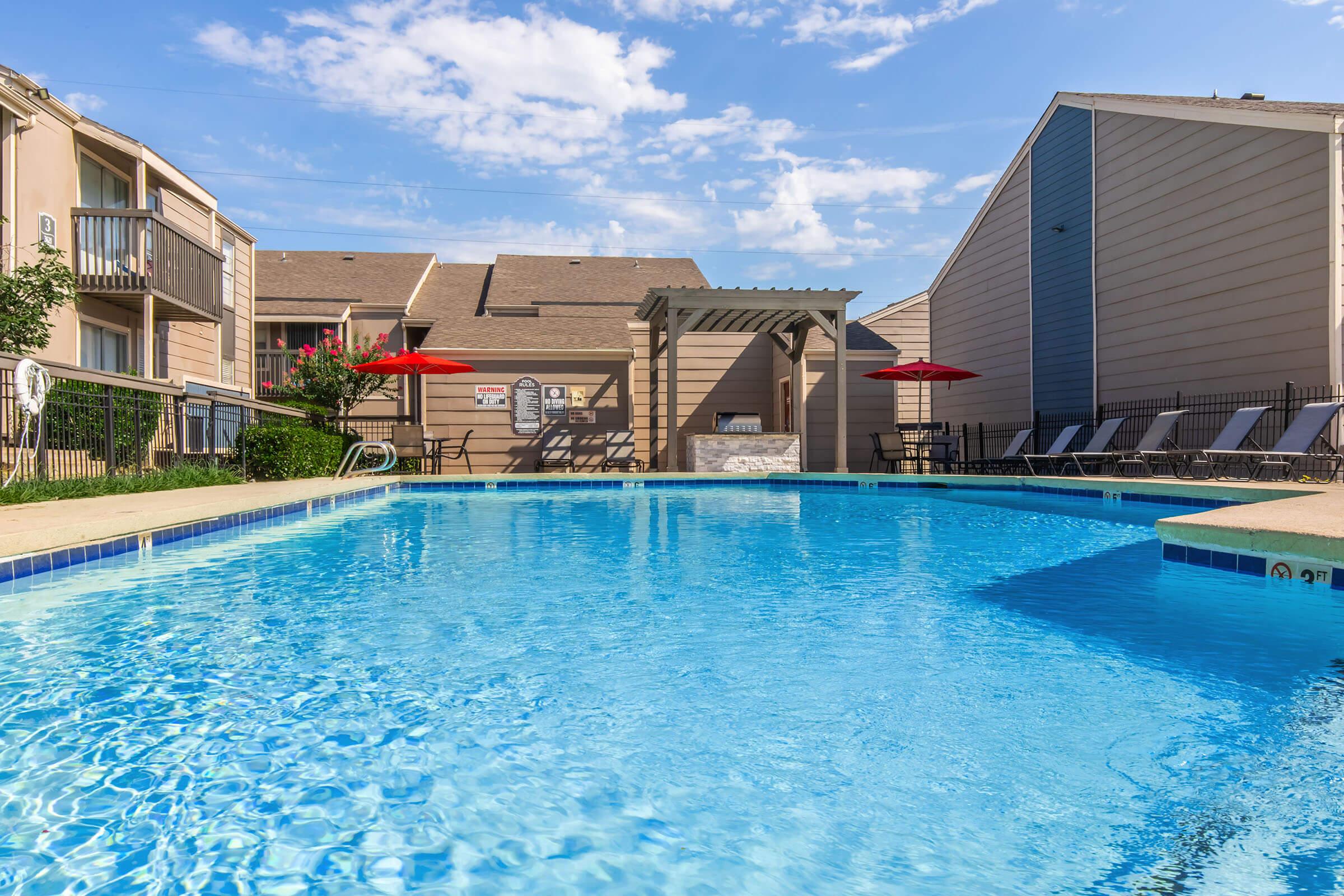 A clear blue swimming pool surrounded by green grass and lounge chairs. There are red umbrellas providing shade near the pool. The area features a pergola and several buildings in the background against a partly cloudy sky.