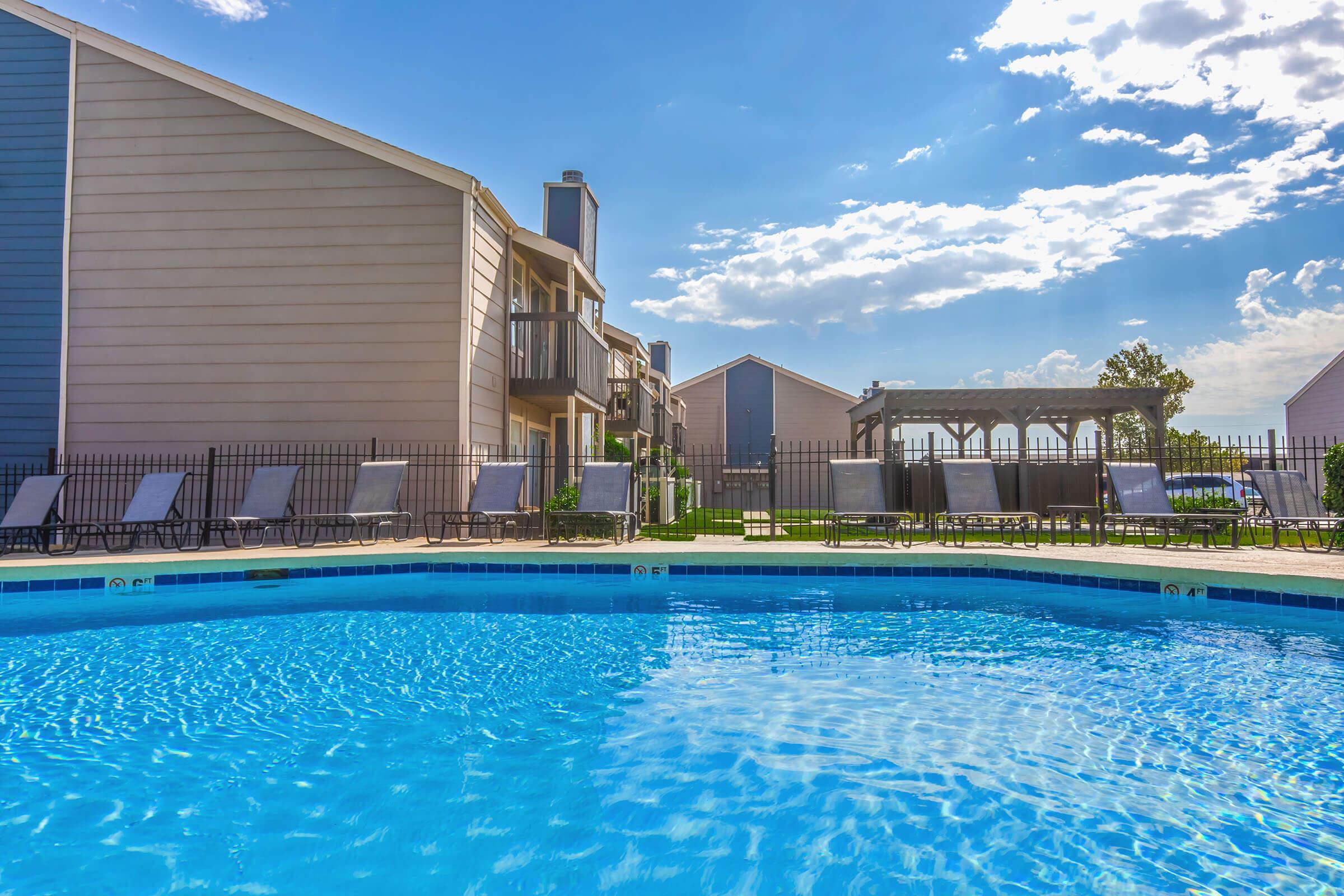 A bright blue swimming pool surrounded by lounge chairs and a fence, with residential buildings in the background. The sky is partly cloudy, and there is a shaded pergola nearby, creating a relaxing outdoor atmosphere.
