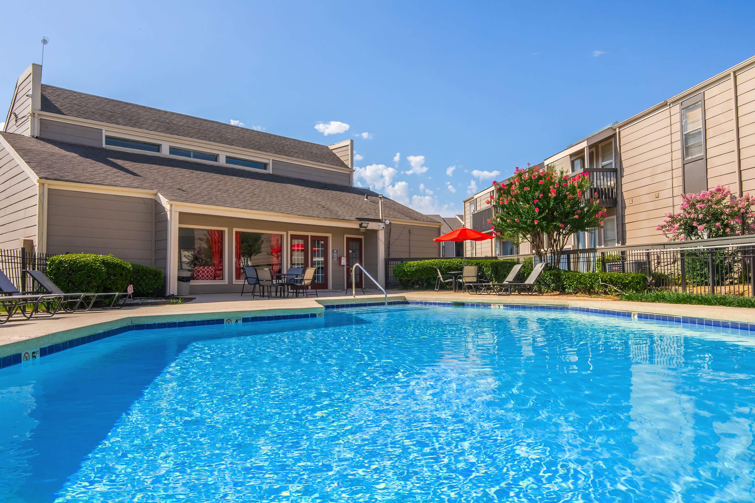 A bright blue swimming pool surrounded by lounge chairs, with a backdrop of a modern apartment building. The pool area features well-maintained landscaping, including trees and shrubs. A patio with red umbrellas is visible, and the sky is clear with a few clouds.