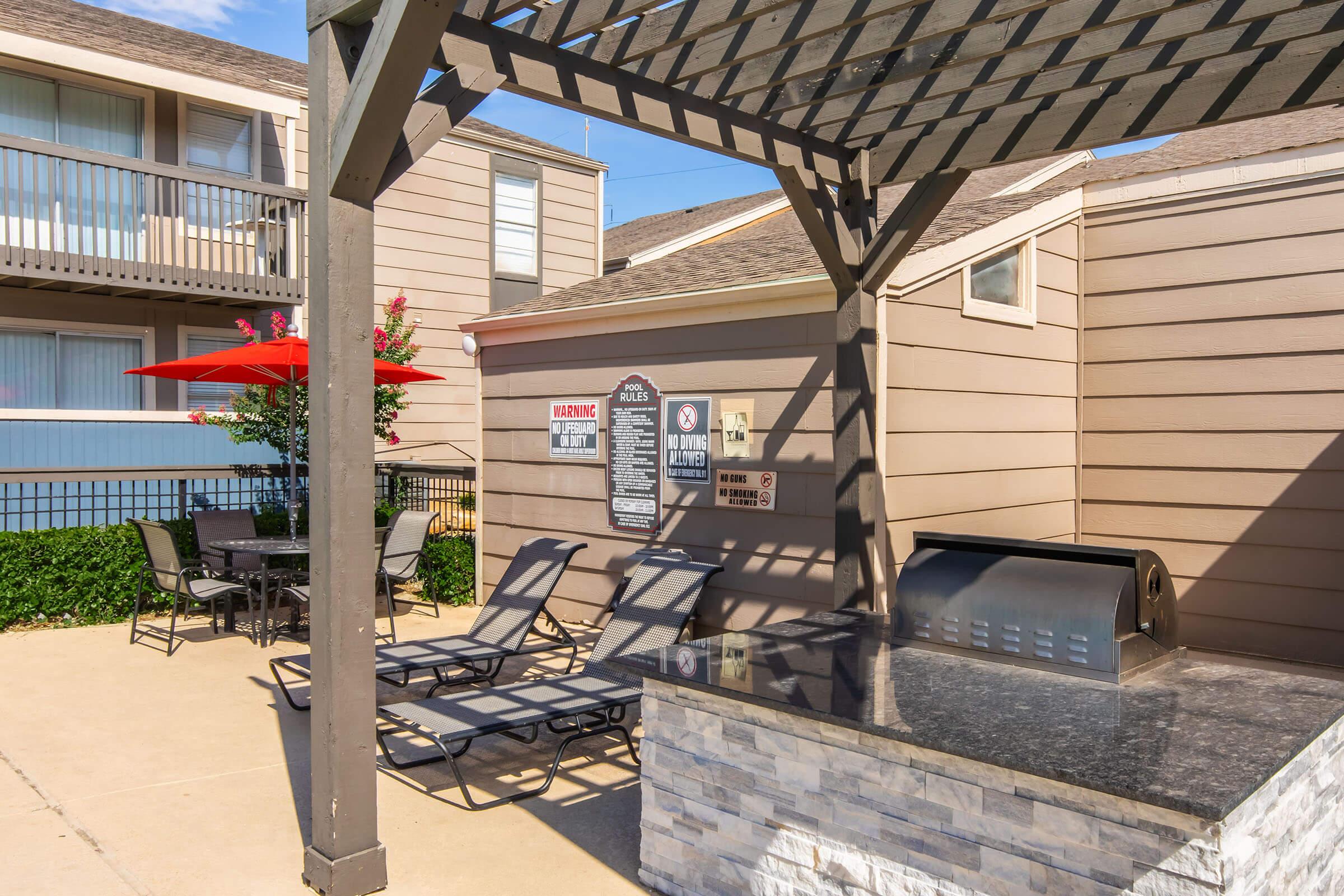 Outdoor recreational area featuring lounge chairs, a stone barbecue grill, and a shaded dining table under an umbrella. The backdrop includes a two-story building with balconies and greenery, creating a relaxed atmosphere suitable for gatherings or relaxation.