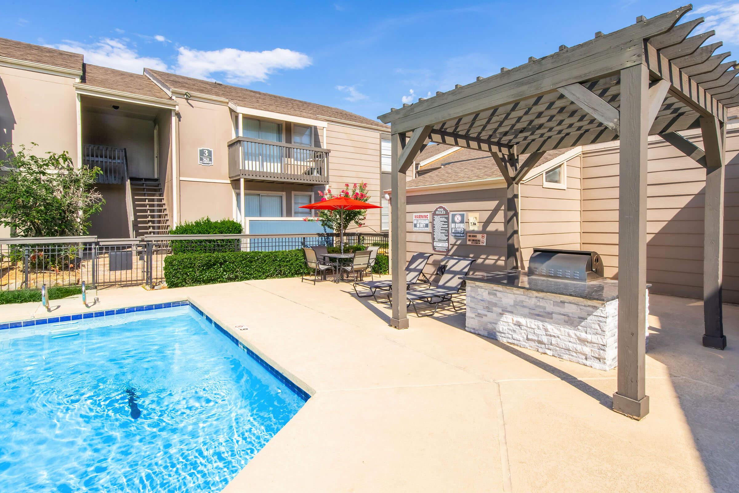 A swimming pool surrounded by lounge chairs and a shaded seating area with a grill. In the background, there are two-story apartment buildings with balconies. A patio umbrella provides shade near the pool, and the sky is clear with a few clouds.