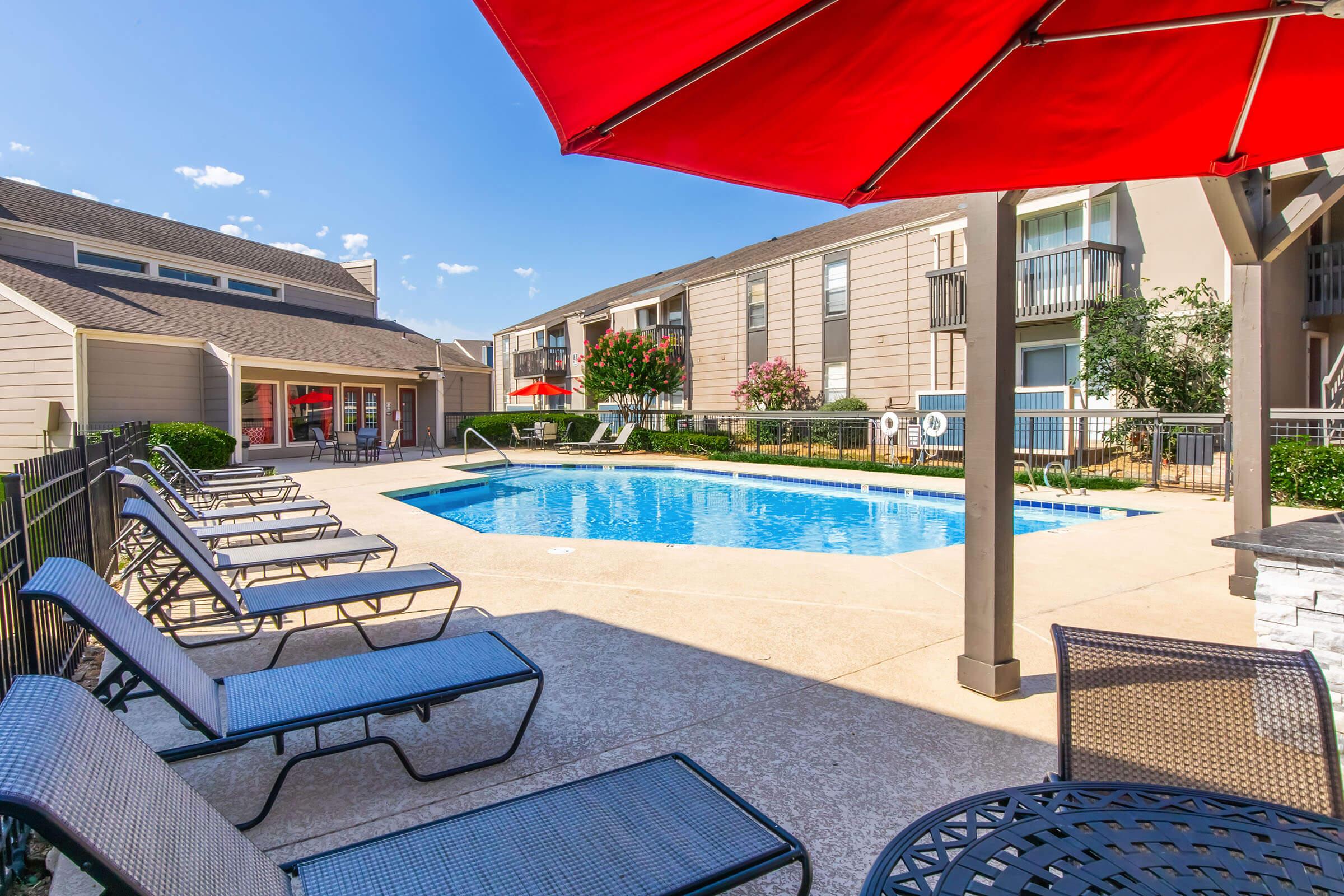 A sunny pool area featuring a clear blue swimming pool surrounded by lounge chairs. Red patio umbrellas provide shade, and well-maintained landscaping adds greenery. Nearby buildings with balconies create a residential vibe. The scene conveys a leisure space ideal for relaxing outdoors.