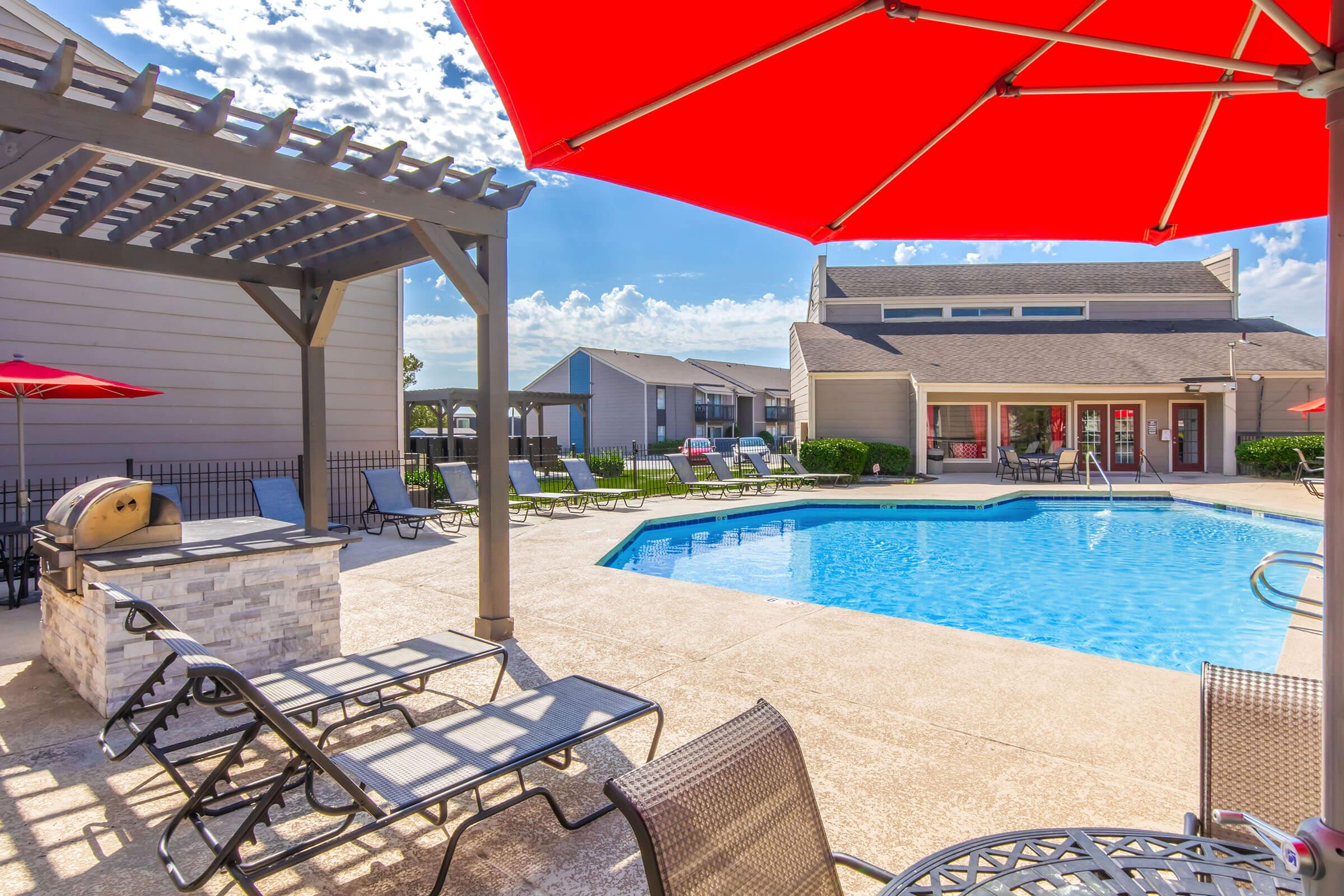 A bright and inviting outdoor pool area featuring a blue swimming pool surrounded by lounge chairs. There are red umbrellas providing shade, and a pergola on one side with a barbecue grill. The sky is partly cloudy, creating a cheerful atmosphere for relaxation and social gatherings.