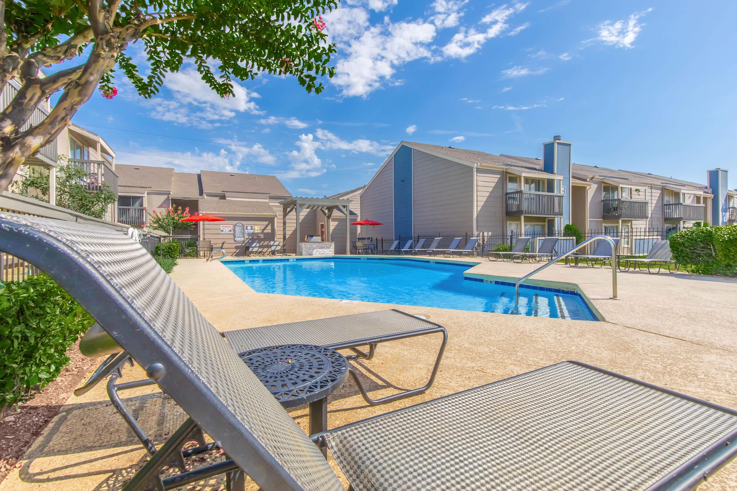 A swimming pool surrounded by lounge chairs, with a vibrant blue sky and a few clouds above. In the background, there are apartment buildings and red umbrellas providing shade near the pool area. Lush greenery and a tree add a touch of nature to the setting.