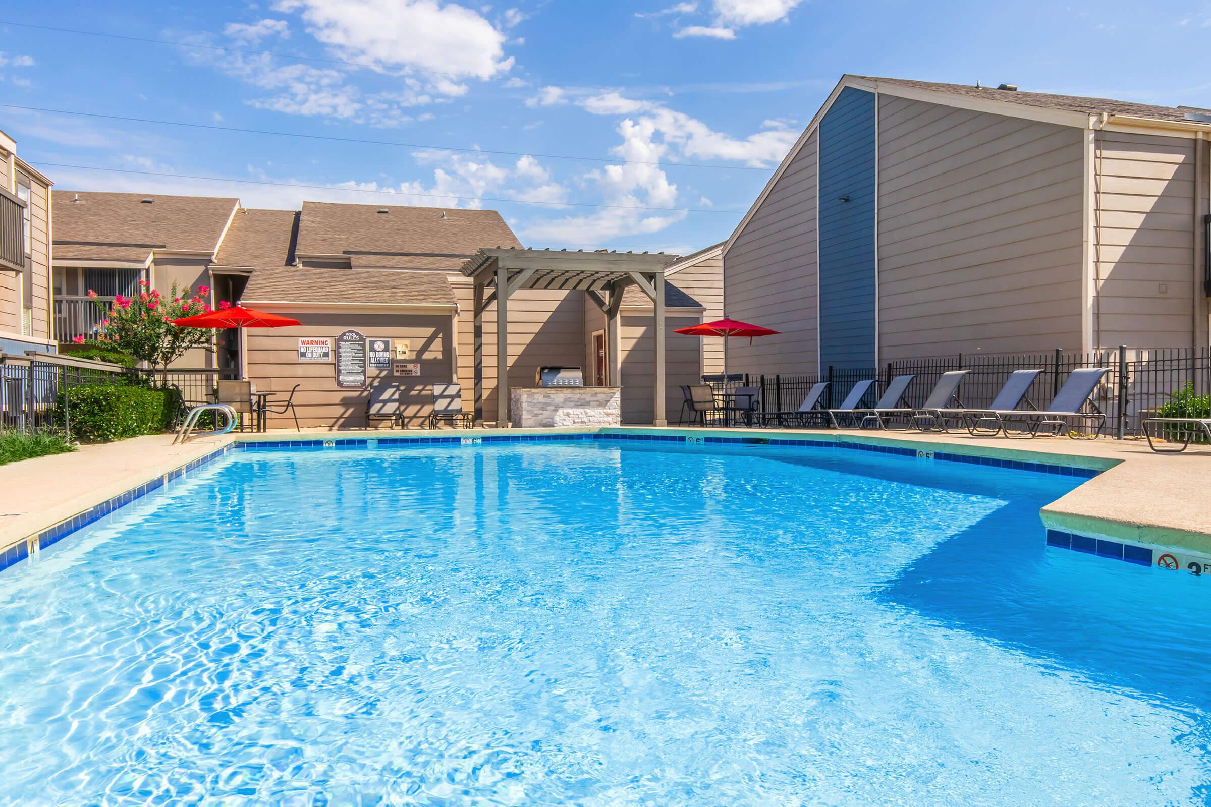 A clear blue swimming pool surrounded by lounge chairs and red umbrellas, with a pergola in the background. The pool is enclosed by a fence, and there are apartment buildings visible in the background under a blue sky with a few clouds.