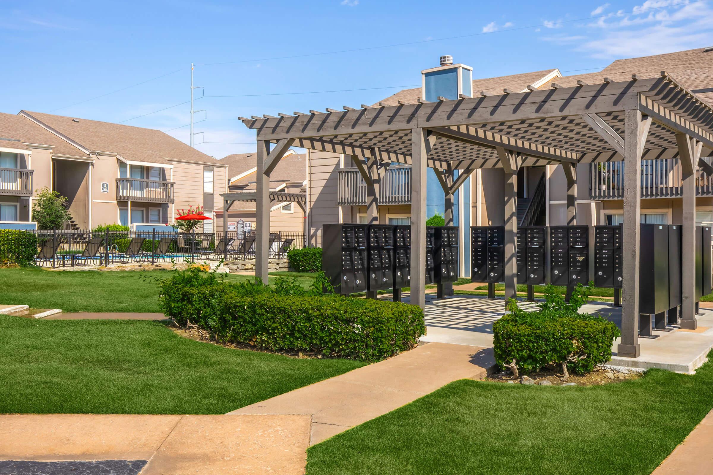 A well-maintained outdoor area featuring a pergola with seating and surrounded by green grass and shrubs. In the background, there are apartment buildings and a clear blue sky. Mailboxes are located near the pergola, adding to the community ambiance.