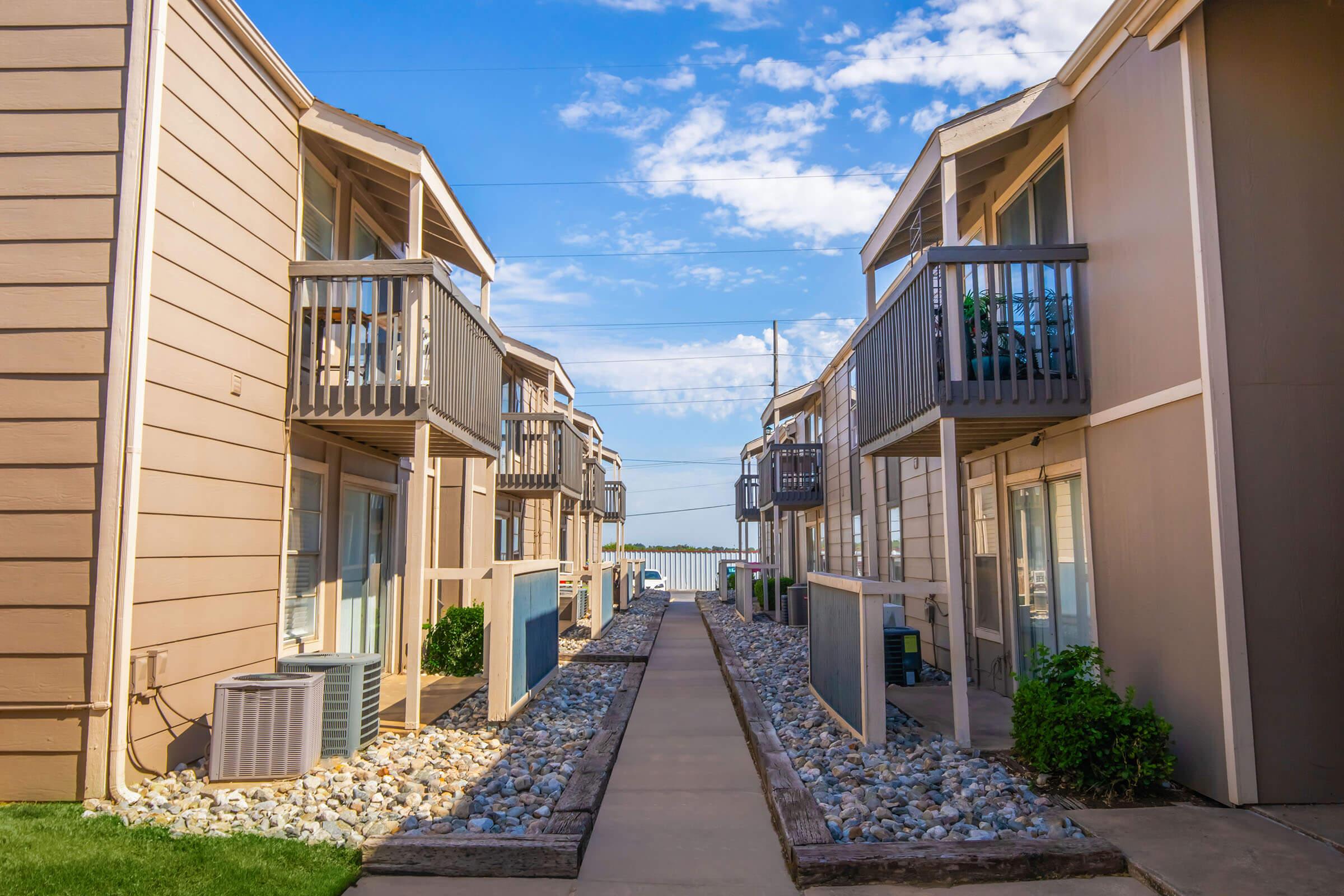 A view of a cozy apartment complex with two-story buildings on either side of a pathway. Each building features a balcony and is surrounded by landscaped areas, including gravel beds. The scene is bright with a clear blue sky and distant water visible at the end of the pathway.