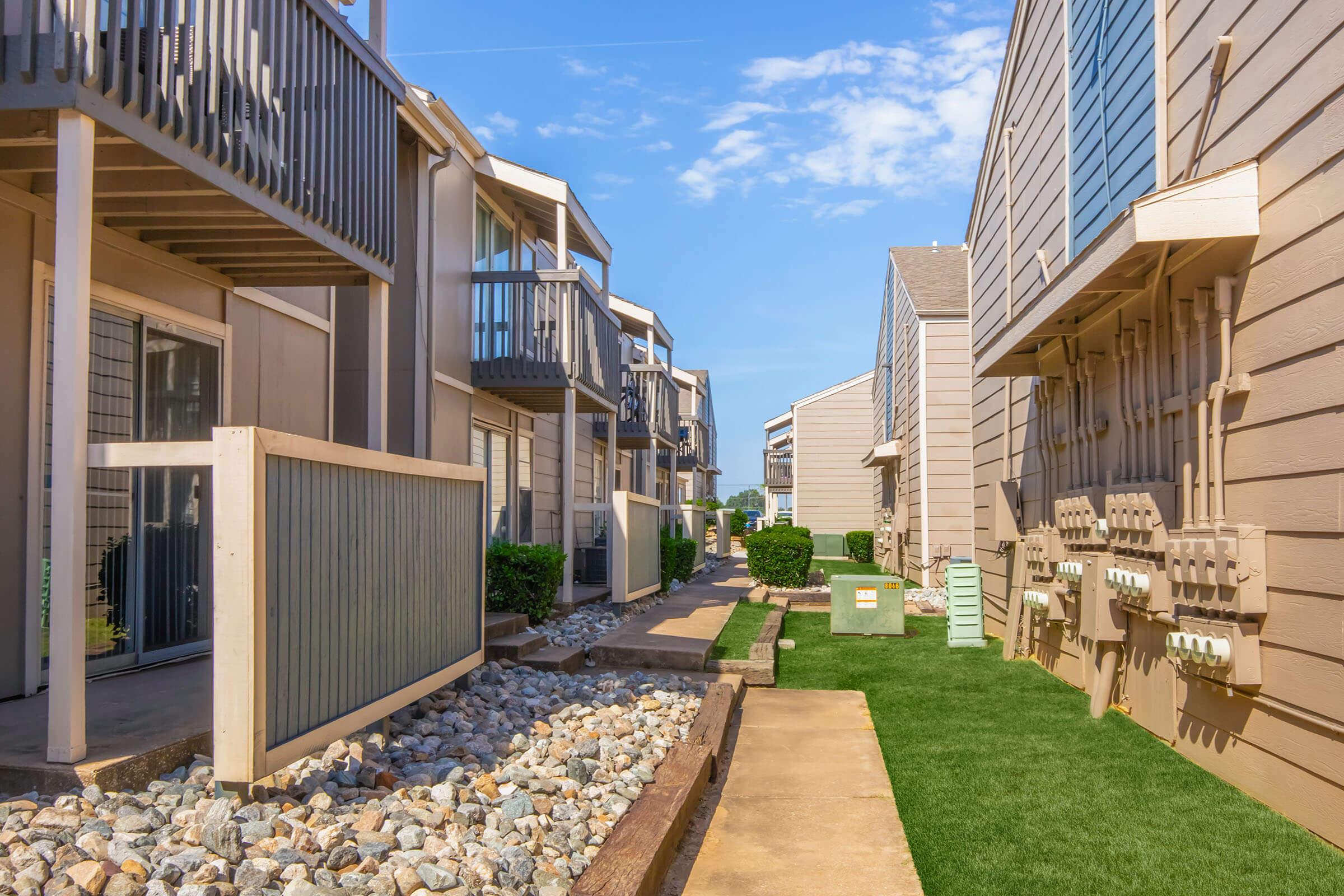 View of a residential area featuring two-story buildings with balconies. A clean walkway lined with rocks and green grass runs between the buildings. Utility boxes are visible on one side, and there are neatly trimmed hedges, creating a tidy and organized appearance. The sky is clear with a few clouds.