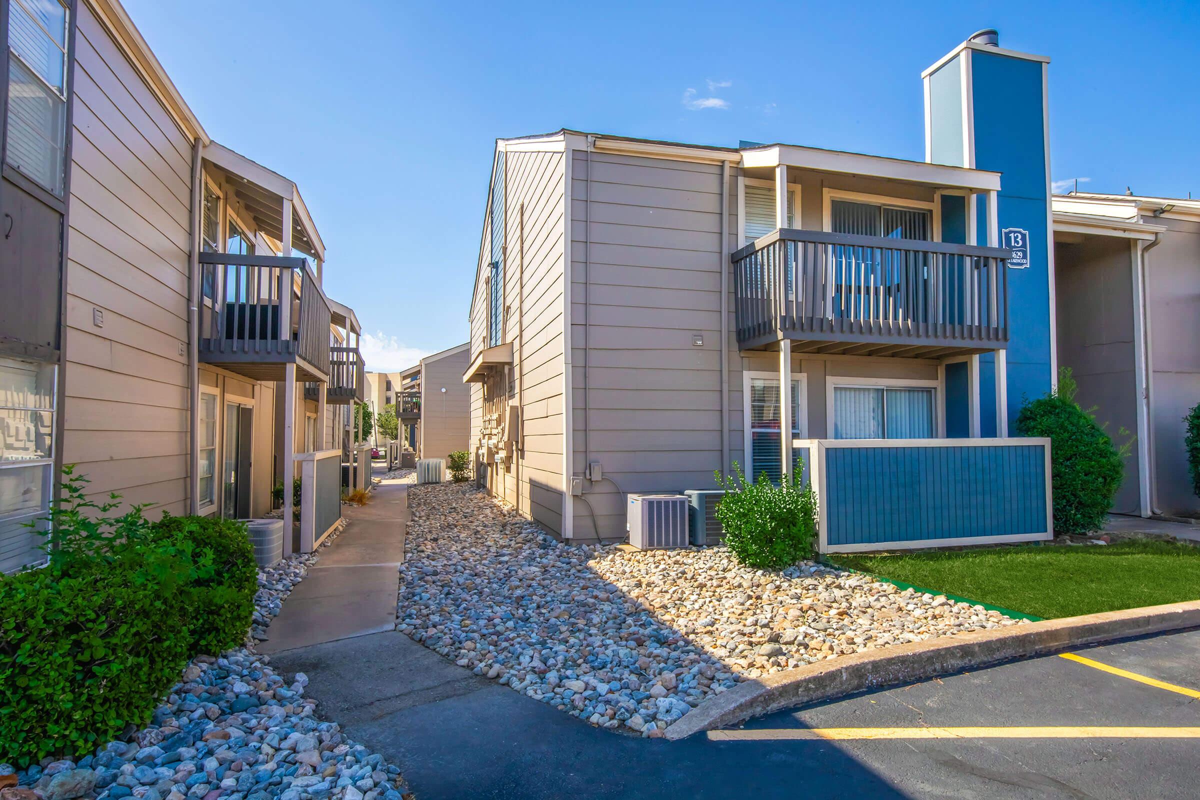 A view of a residential complex featuring two-story buildings with balconies, a walkway lined with rocks, and green landscaping. Bright sunlight illuminates the scene, highlighting the exterior design and neat surroundings.