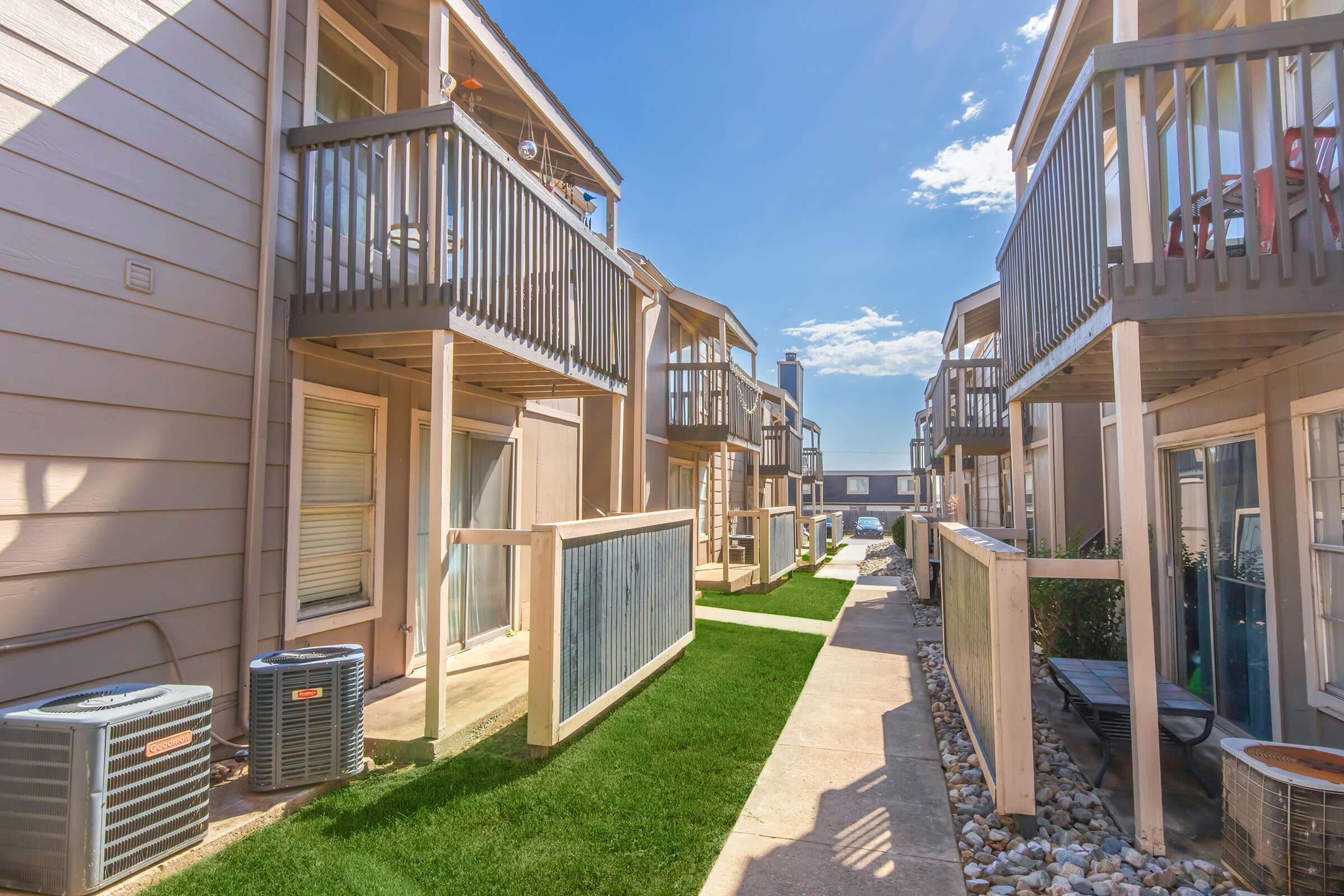 A narrow pathway between two rows of multifamily buildings, featuring balconies and air conditioning units. The walkway is lined with manicured grass and decorative stones, under a bright blue sky with a few clouds. The scene conveys a pleasant, well-maintained outdoor living space.