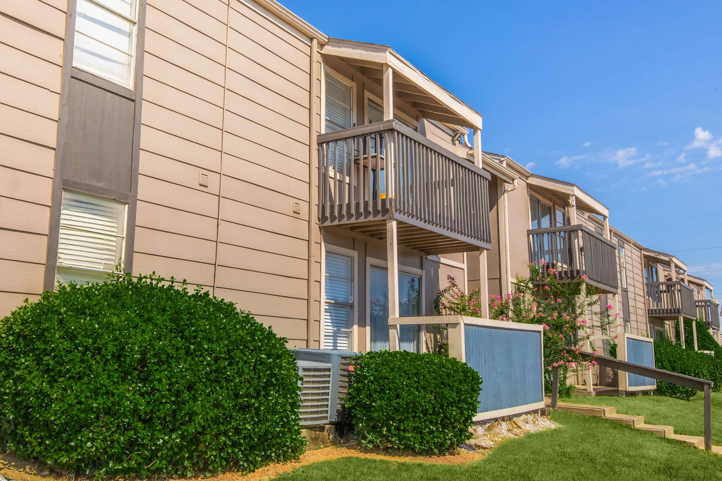 A row of apartment buildings with light brown wood siding. Each unit has a small balcony with railings. Lush green shrubbery is planted in front, and there's a well-maintained lawn. The sky is clear and blue, creating a bright and inviting atmosphere.