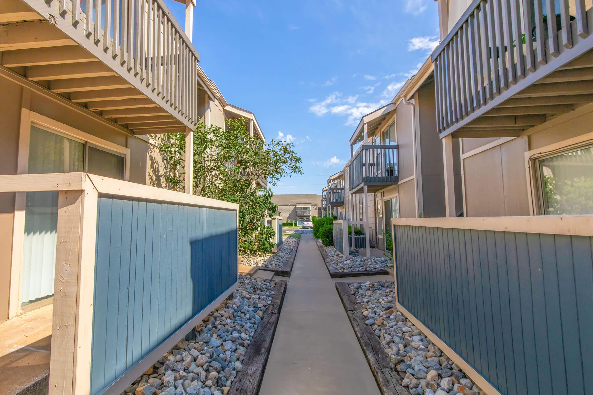 A serene pathway between two sets of multi-unit buildings, flanked by blue and beige wooden fences. The path is lined with pebbles and greenery, under a bright blue sky with a few clouds. The scene conveys a peaceful, well-maintained outdoor environment.