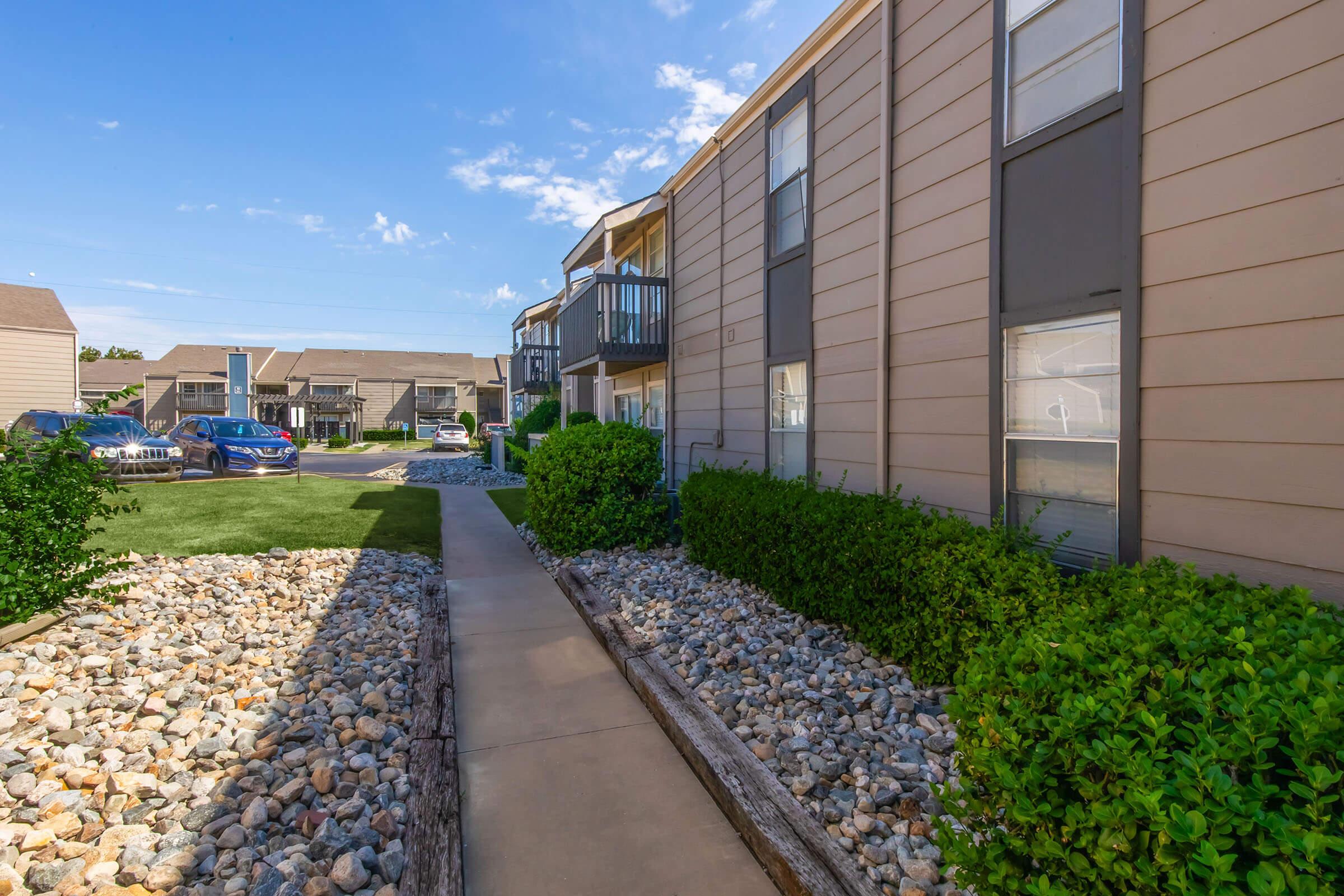 A walkway lined with rocks and shrubs leads to a series of apartment buildings. The buildings have beige siding with balconies and large windows. The sky is clear with some clouds, and there are cars parked nearby in the background. The area is well-maintained and appears inviting.