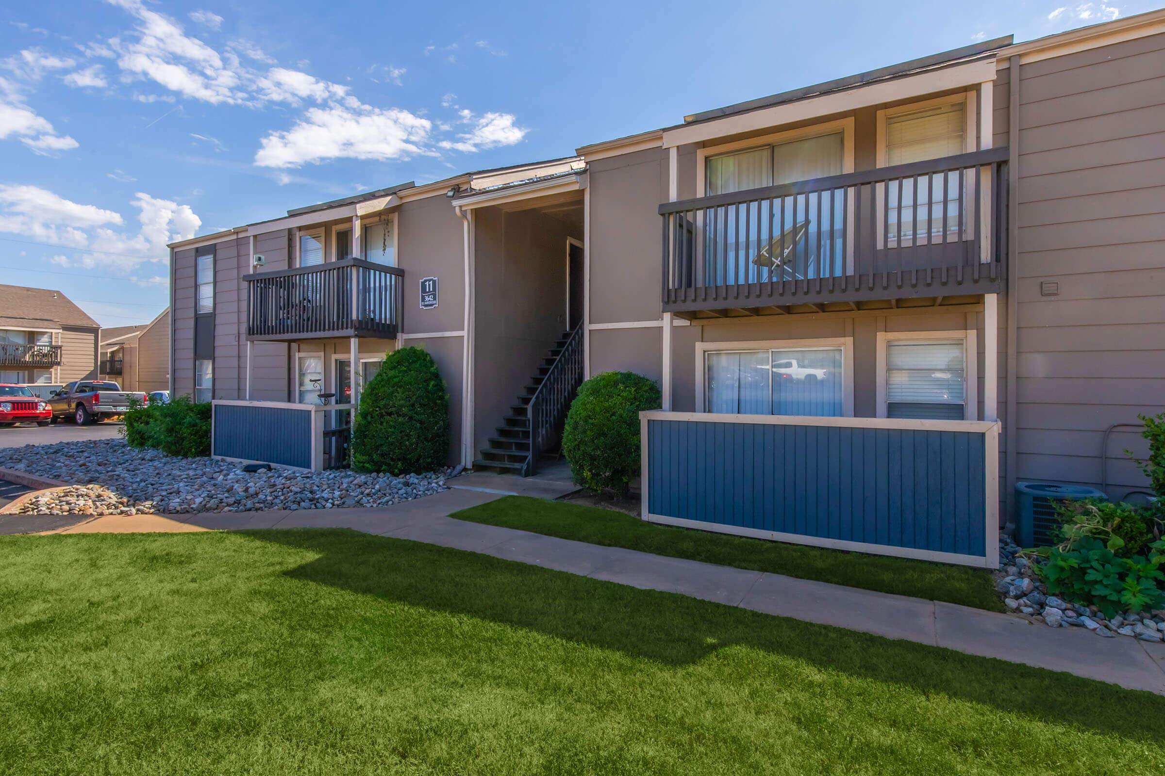 Two-story apartment building with balconies, nestled between green lawns and landscaped bushes. A pathway leads along the front, with a glimpse of parked cars in the background. The sky is partly cloudy, enhancing the suburban atmosphere.