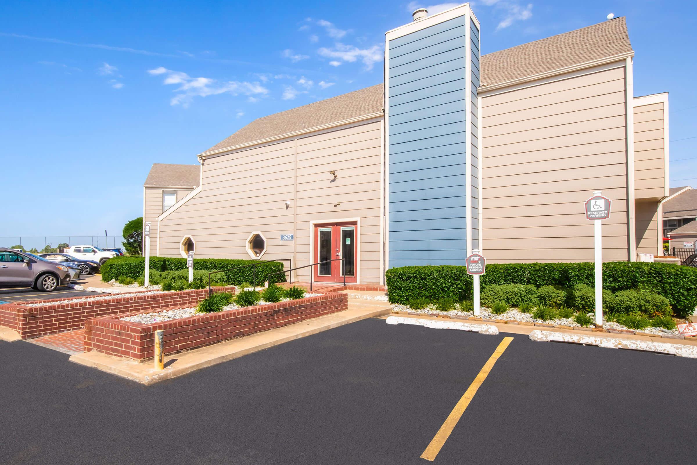 Exterior view of a modern building with a combination of beige and blue siding. The entrance features glass doors, surrounded by neatly trimmed hedges and landscaping. A parking lot is visible in front, with designated parking signs and a clear blue sky in the background.