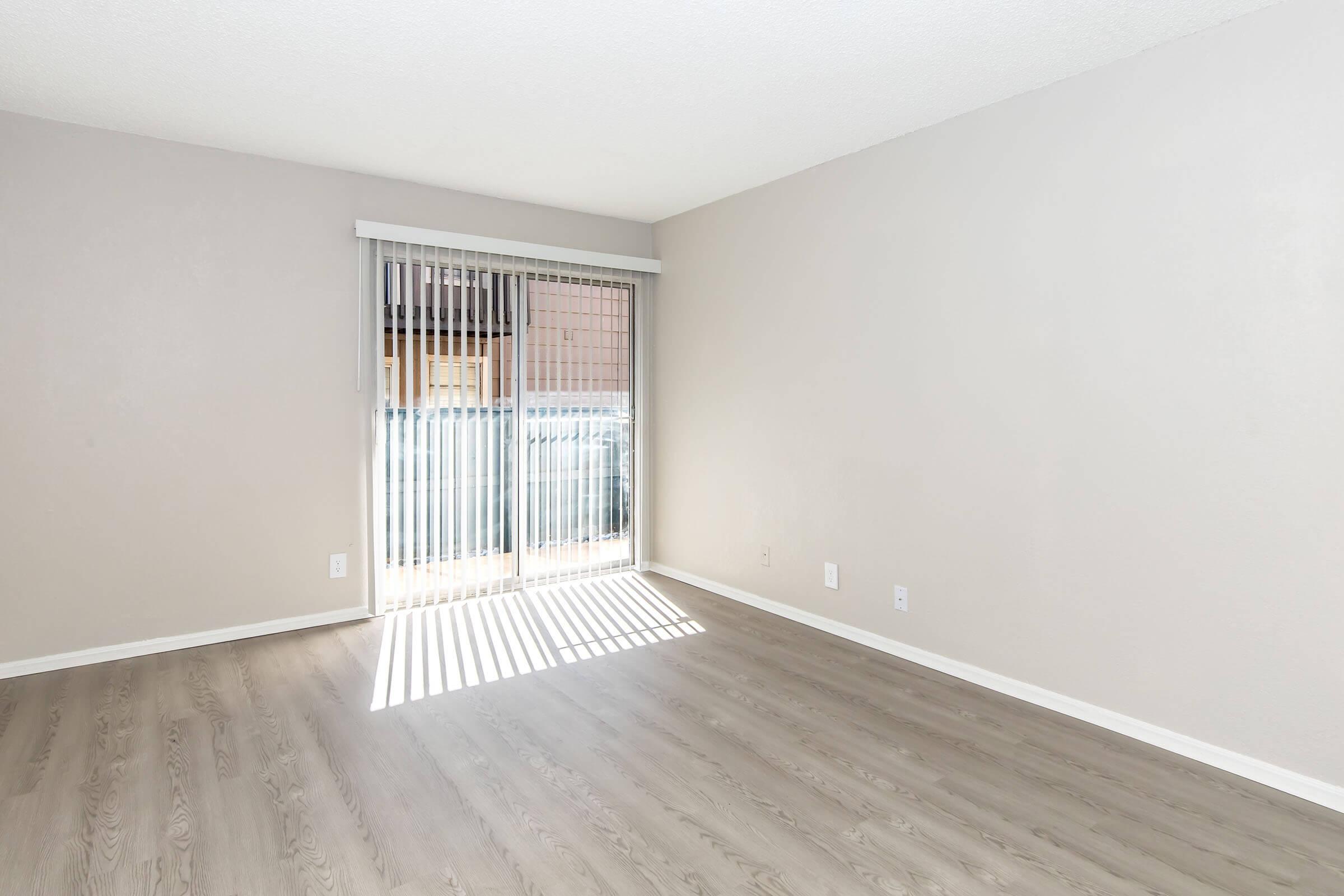 A vacant room with light brown laminate flooring and light gray walls. Large sliding glass doors with vertical blinds allow natural light to enter, casting shadows on the floor. The room has a clean, minimalist aesthetic with no furniture or decorations.