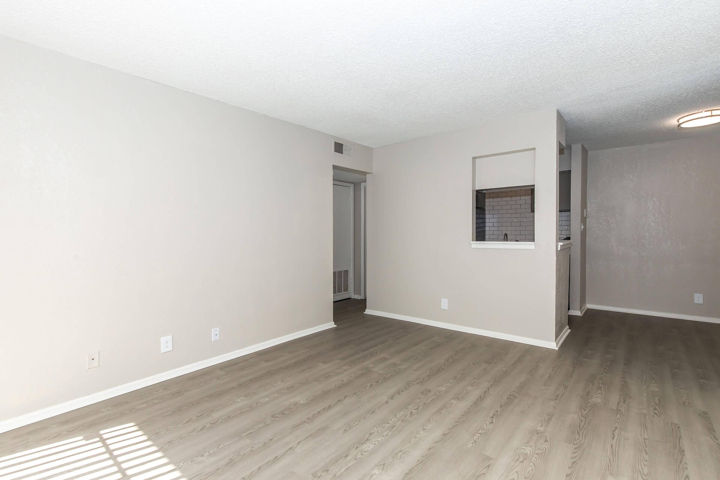 Empty living room with light-colored walls and laminate flooring. A doorway leads to another room, and a partially visible kitchen area can be seen in the background. The space is bright and open, emphasizing a clean, modern aesthetic with minimal furnishings.