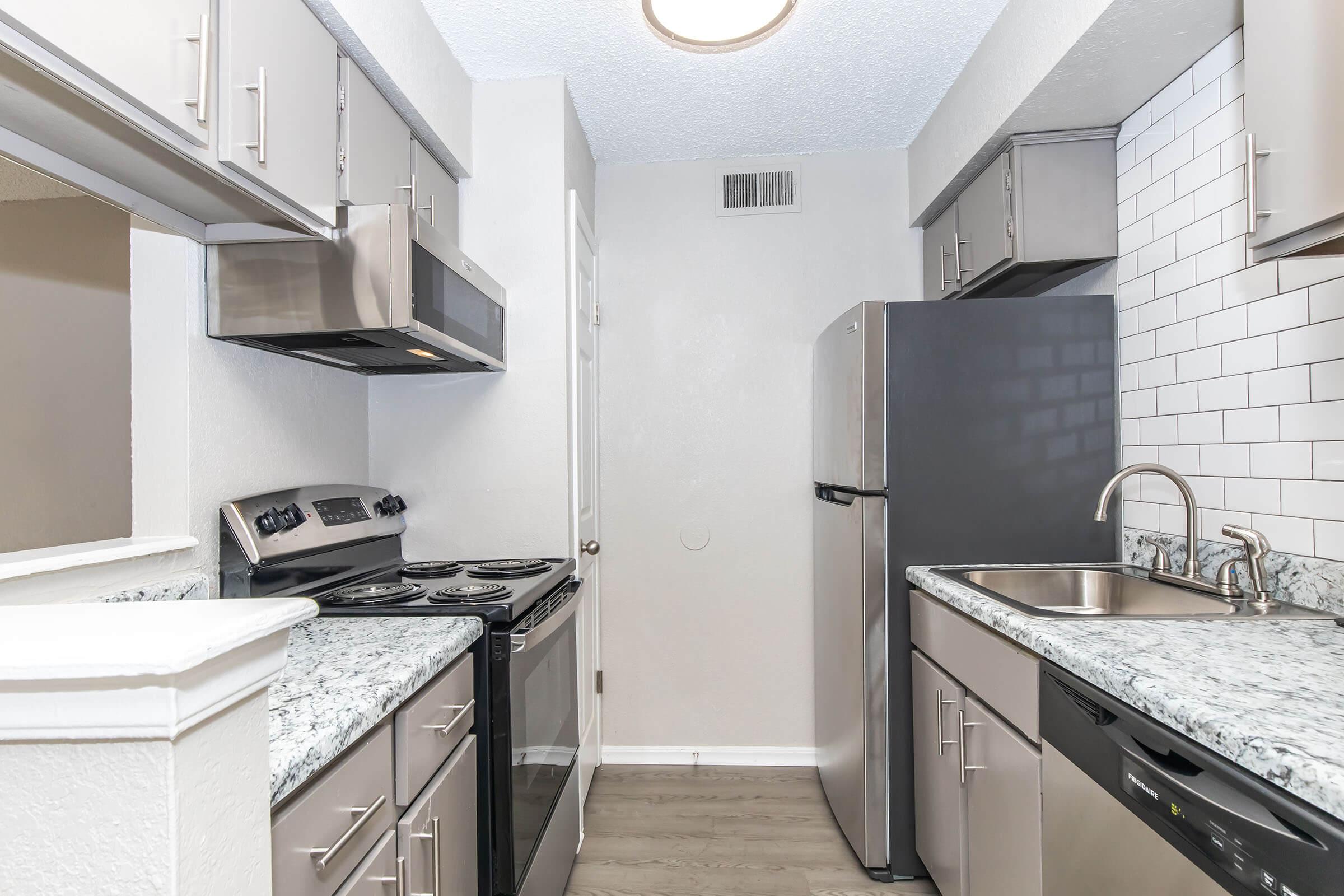 A modern kitchen featuring stainless steel appliances, including an oven, stovetop, microwave, refrigerator, and dishwasher. The cabinets are sleek with a neutral color scheme, complemented by granite countertops and a white tiled backsplash. The flooring is light and contemporary.