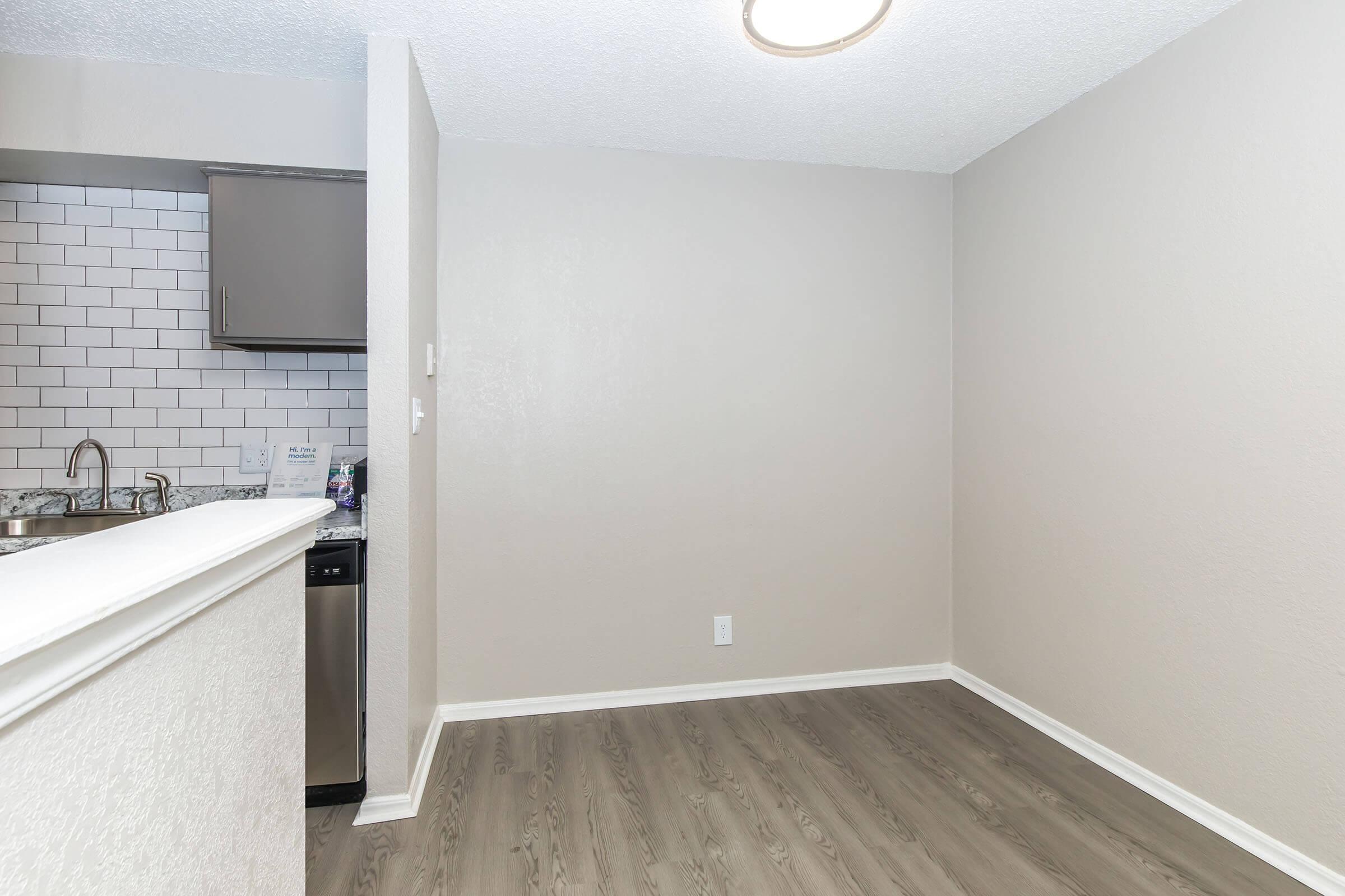 A modern kitchen area with a gray color scheme. There is a minimalistic wall with light-colored paint, a ceiling light fixture, and a partial view of a tiled kitchen counter with a stainless steel sink and dishwasher. The floor is finished with light wood-like flooring.