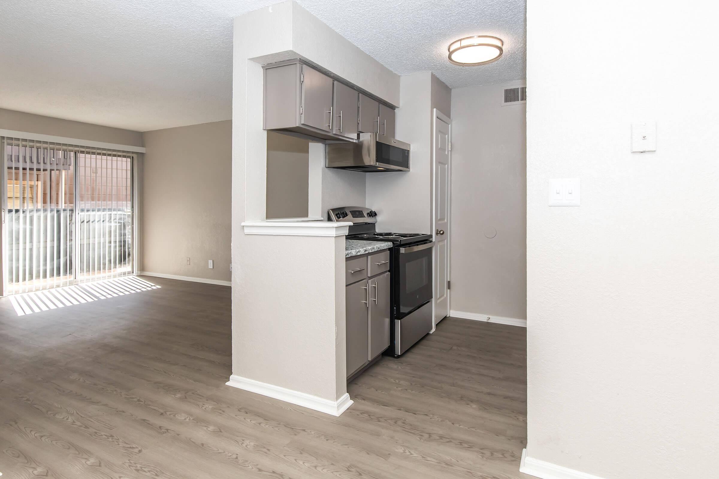 Modern kitchen and living area in a bright, open-plan layout. The kitchen features gray cabinets, a stove, and a hood, while the living area is illuminated by natural light from sliding glass doors leading to an outdoor space. Neutral wall colors and laminate flooring create a clean, inviting atmosphere.