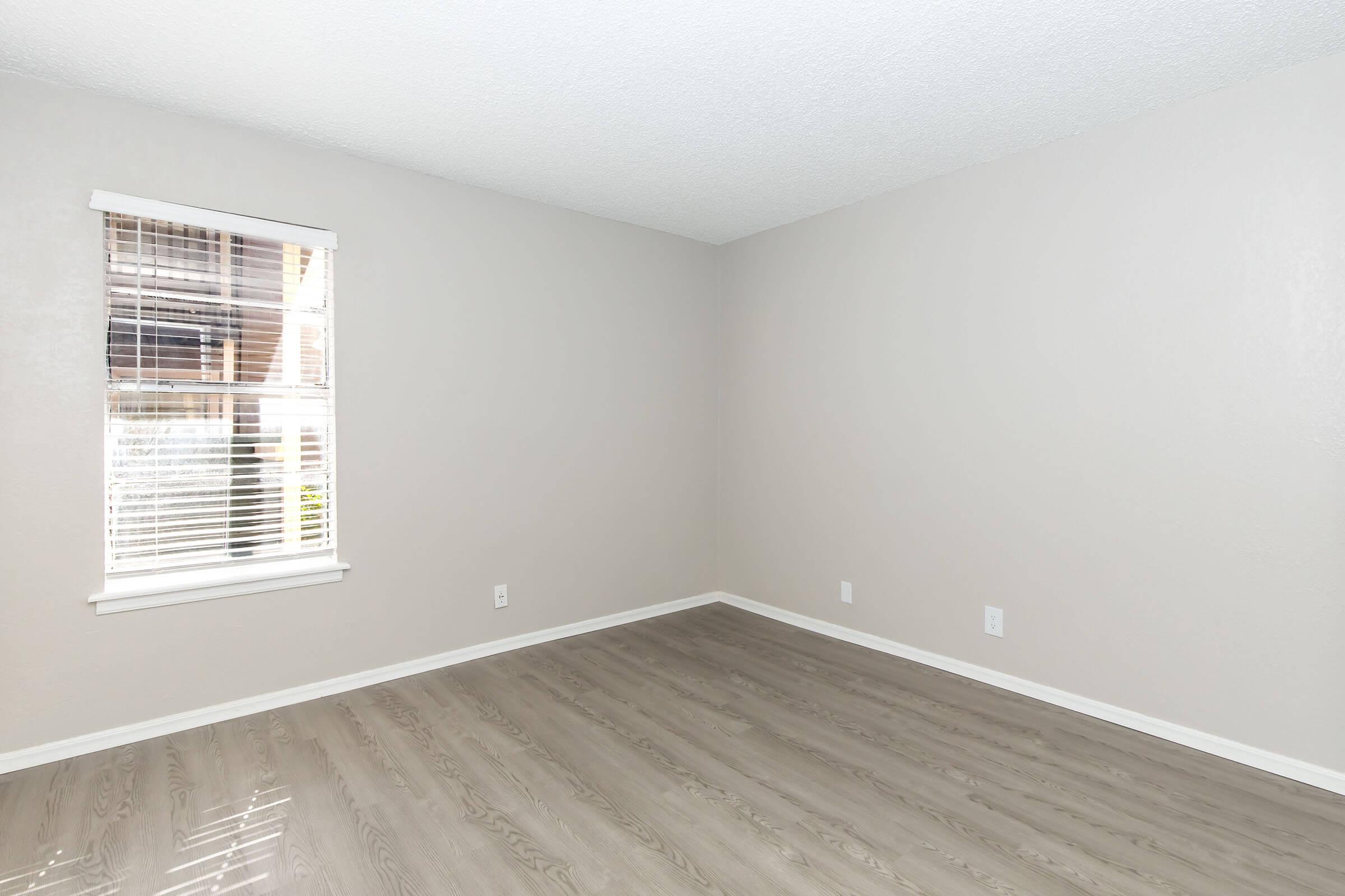 Empty room with light beige walls and light-colored wood flooring. A window with white blinds lets in natural light. The room is sparsely decorated with no furniture, creating a clean and open space.
