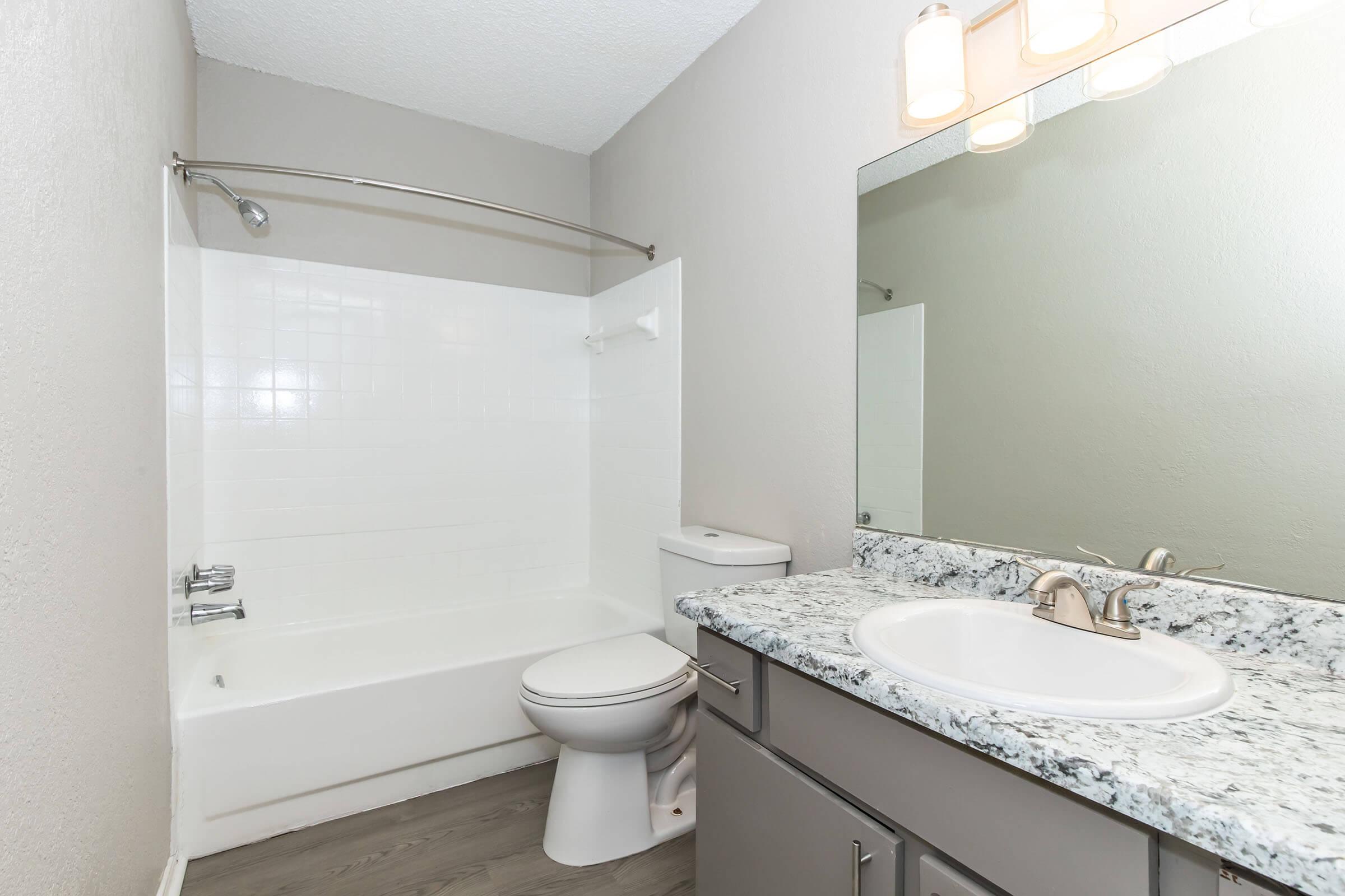 Modern bathroom with a white bathtub and shower combination, a sleek toilet, and a granite countertop sink with a single faucet. A rectangular mirror is positioned above the sink, and there is a shower rod on the wall. The walls are painted in a light gray color, and the floor features a wood-like design.