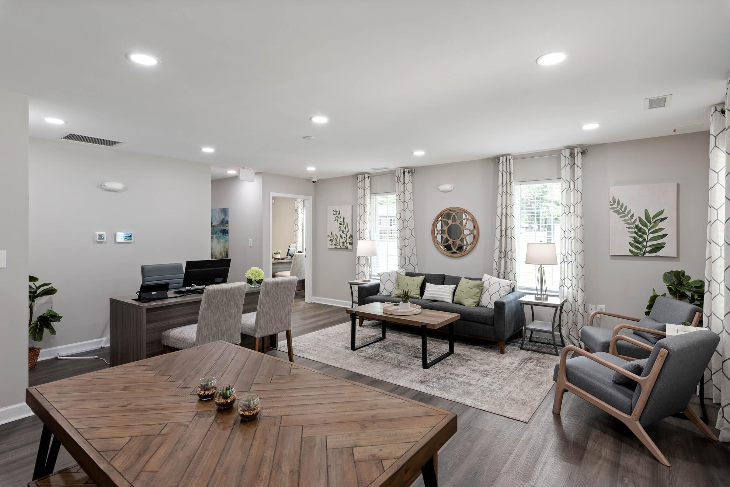 A modern living room with a cozy seating area, featuring a gray sofa, wooden coffee table, and stylish armchairs. Potted plants add greenery, while artwork of plants decorates the walls. Soft curtains frame the windows, and a well-lit home office space is visible in the background.