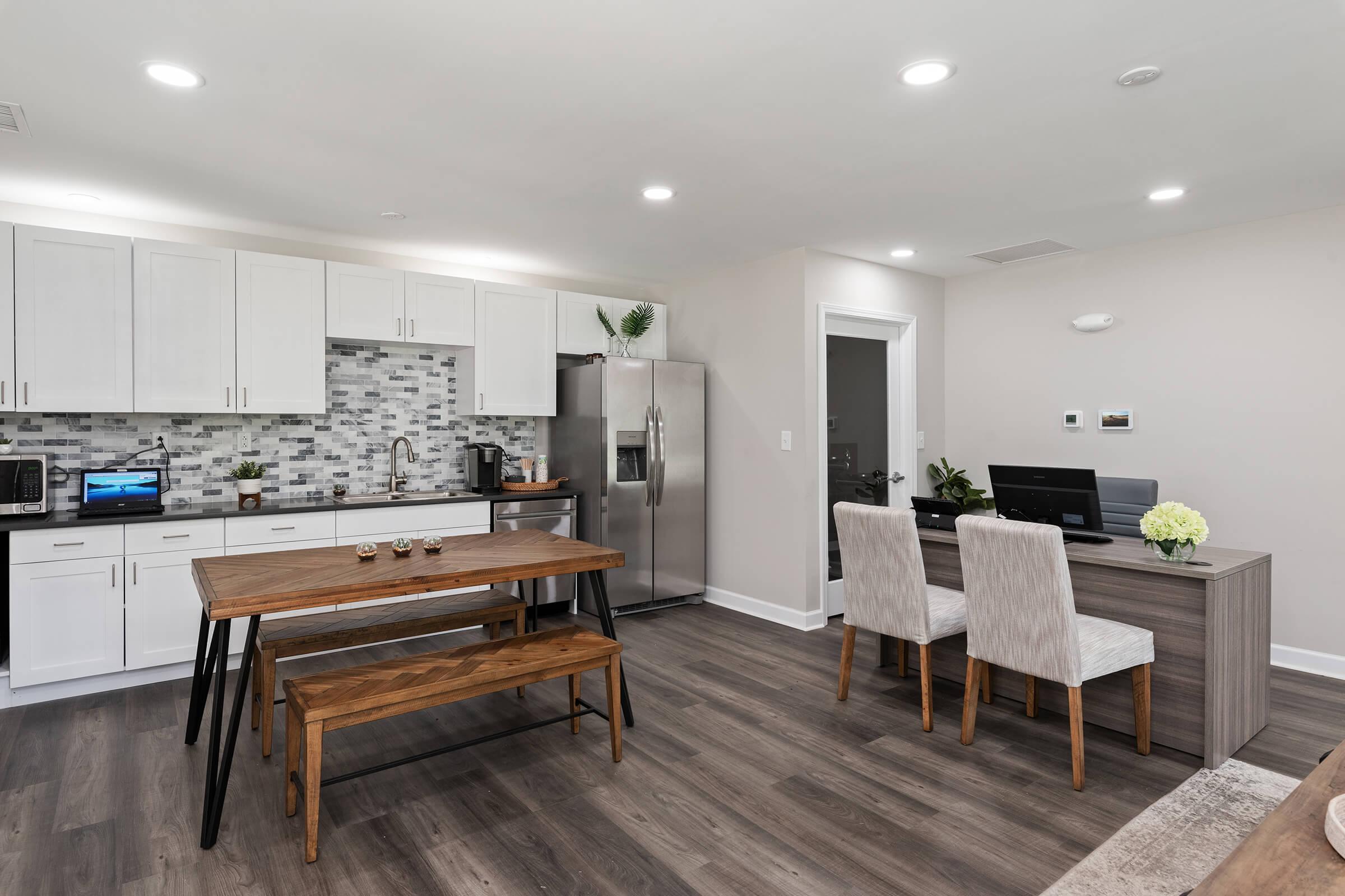 Modern kitchen and dining area featuring white cabinetry, a gray tiled backsplash, a stainless steel refrigerator, and a wooden dining table with benches. A work desk with a computer is situated next to the kitchen, and the flooring is a warm wood tone, creating a cozy and inviting atmosphere.