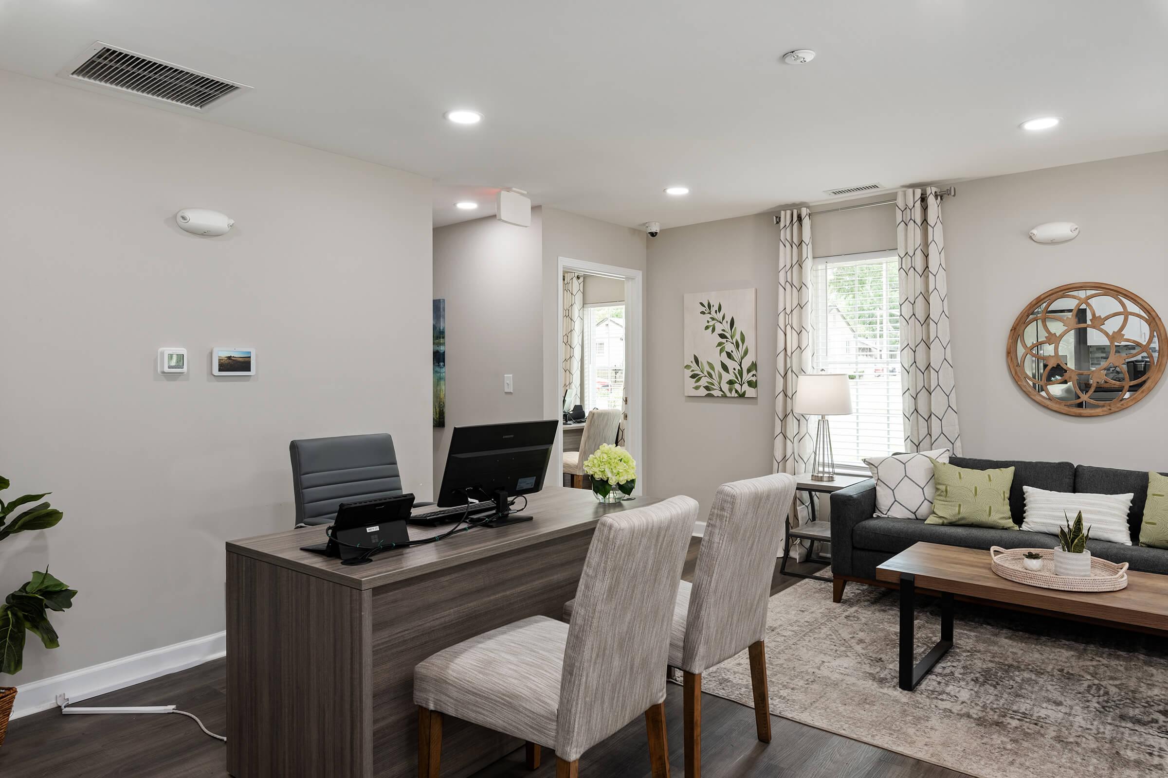 A modern reception area featuring a wooden reception desk with a computer, two upholstered chairs, and decorative plants. The walls are painted in neutral tones, and large windows bring in natural light. A cozy seating area includes a gray sofa, a coffee table, and a round wall mirror.