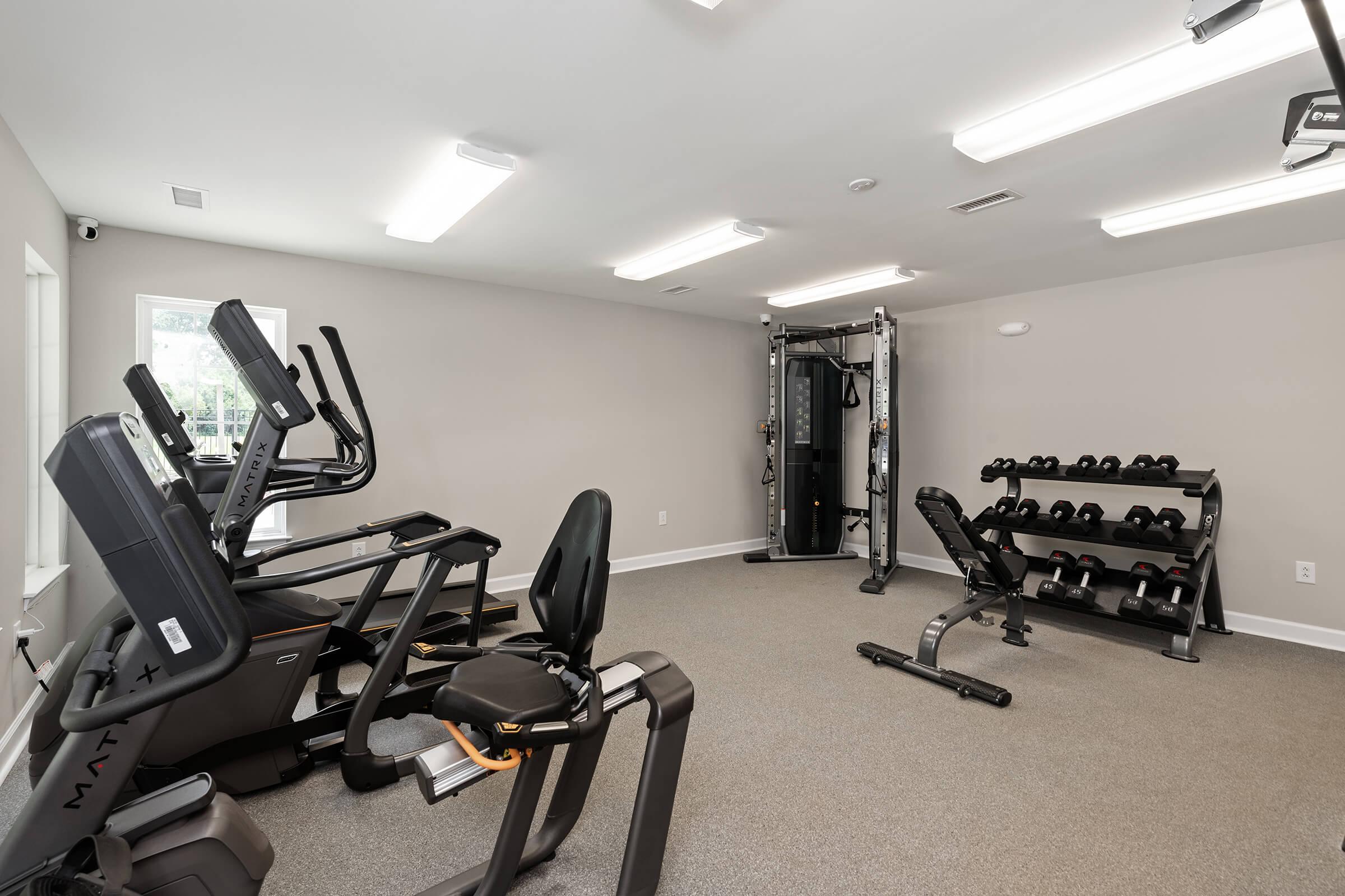A modern fitness room featuring various gym equipment, including treadmills, exercise bikes, and a weight bench with dumbbells stacked on a rack. The walls are painted in a neutral color, and the room is well-lit with overhead LED lighting. The flooring is carpeted for comfort.