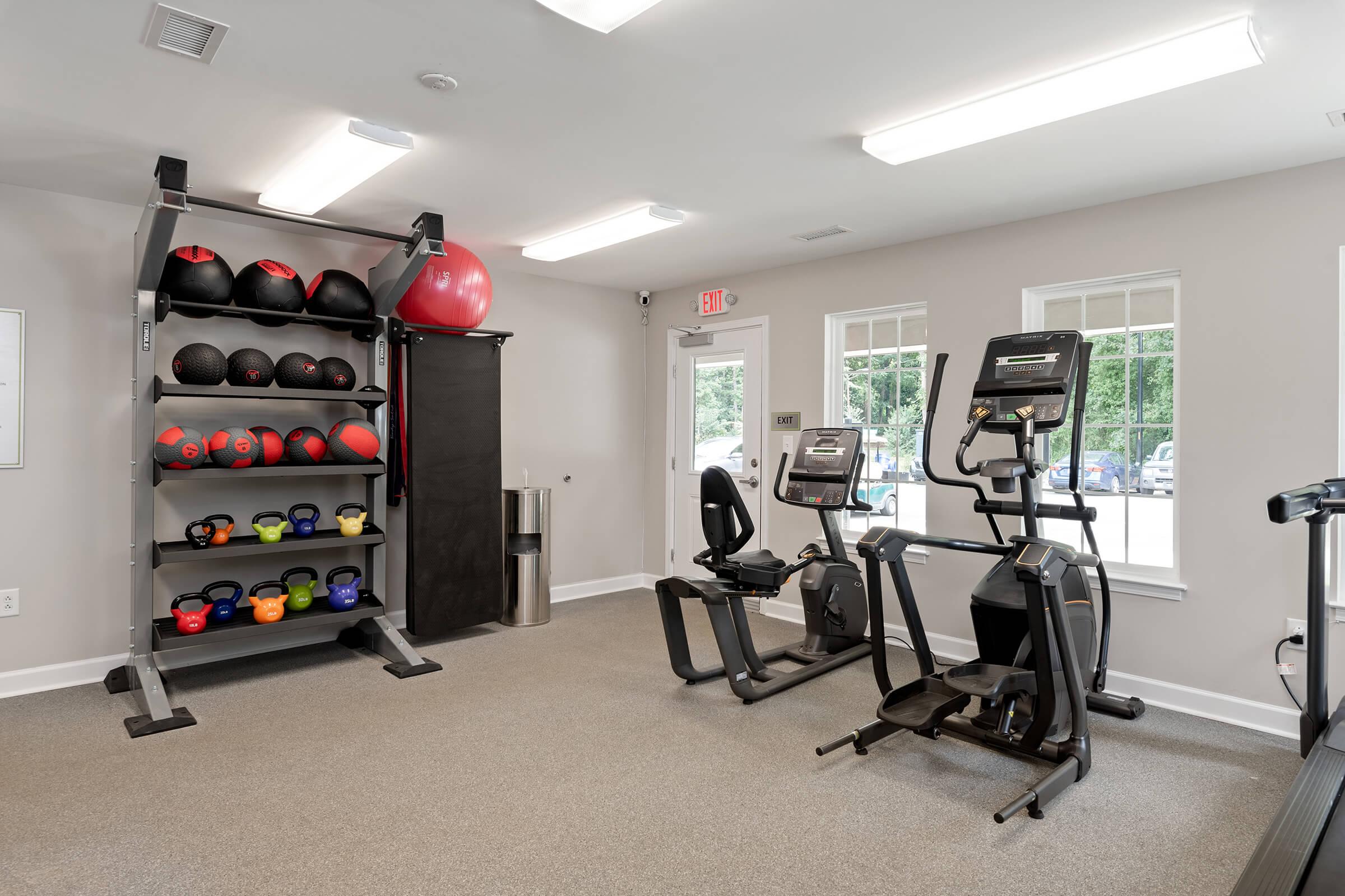 A fitness room featuring a rack of colorful exercise balls and kettlebells, alongside two pieces of exercise equipment: a recumbent bike and an elliptical machine. Large windows provide natural light, and there is a waste bin in the corner. The space is well-lit and inviting for workouts.