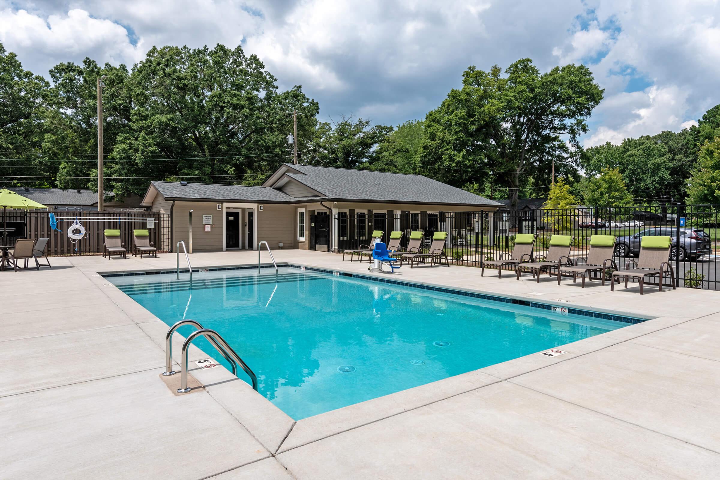 A bright outdoor swimming pool surrounded by lounge chairs, with a person sitting by the poolside. The area is well-maintained, featuring greenery and a nearby building. The sky is partly cloudy, creating a pleasant atmosphere for relaxation and leisure.