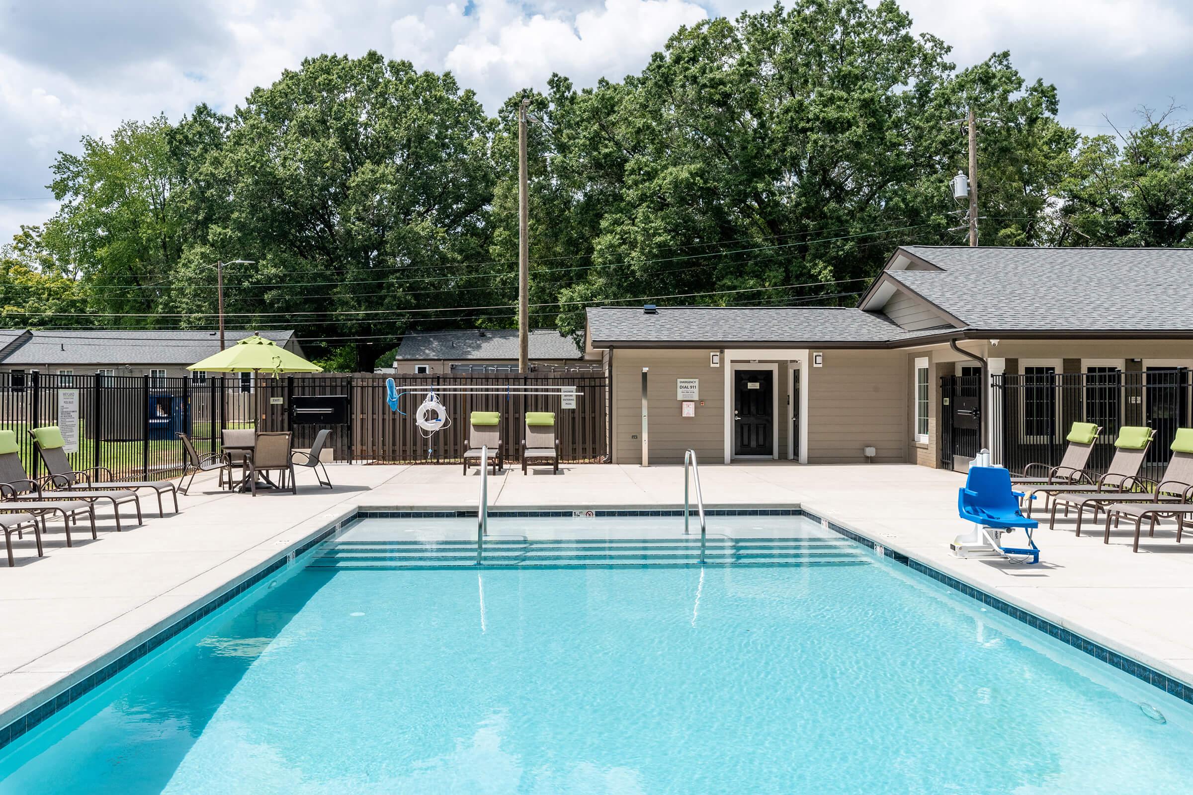 A clear swimming pool surrounded by lounge chairs and umbrellas, with a poolside accessible chair. In the background, there are trees and a building with a door leading inside. The scene is bright and inviting, perfect for relaxation and recreation.