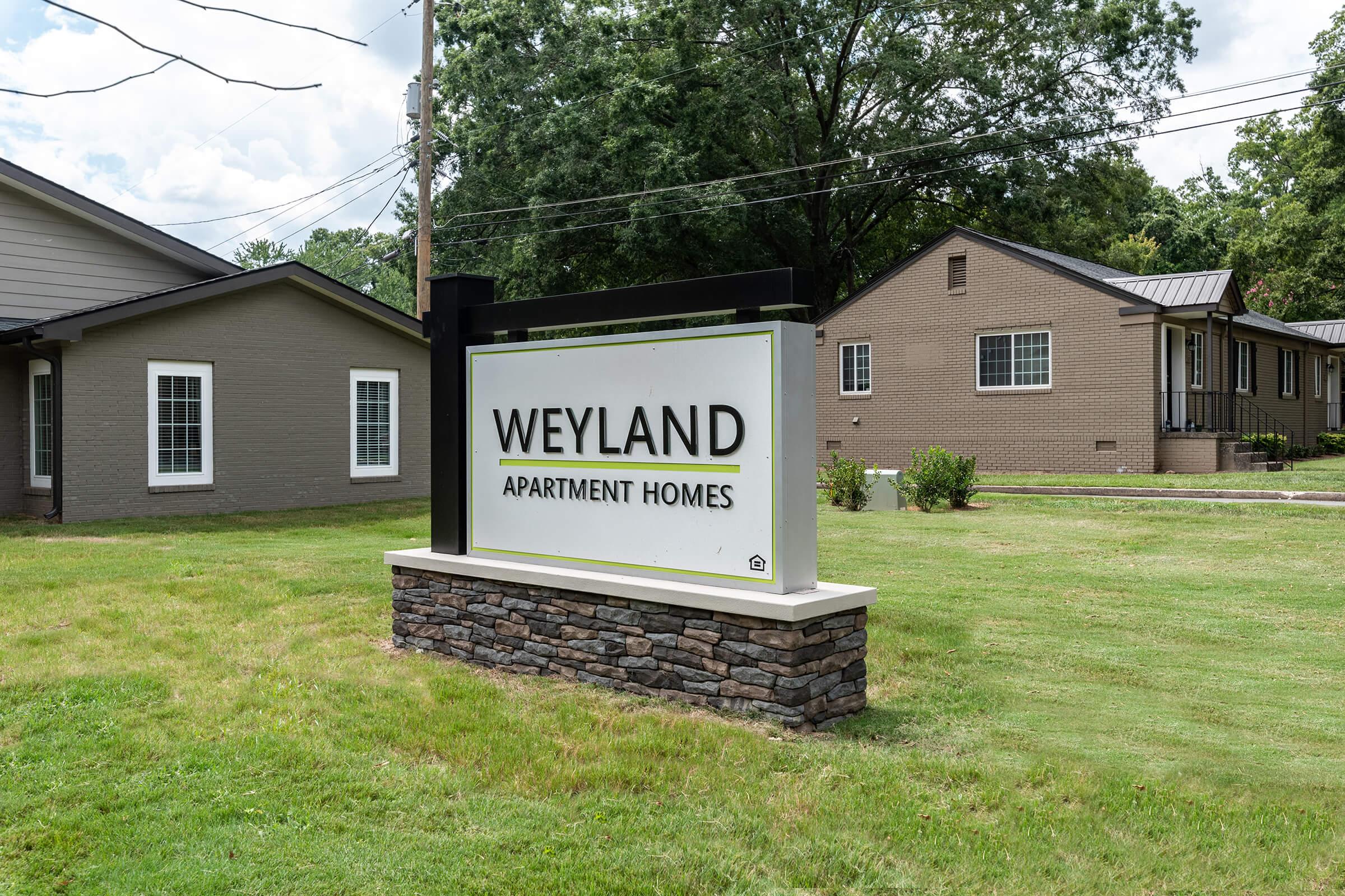 Sign for Weyland Apartment Homes, featuring the name prominently displayed on a white background. The sign is supported by a stone base and is situated in a grassy area with several residential buildings visible in the background. Trees provide a natural backdrop.