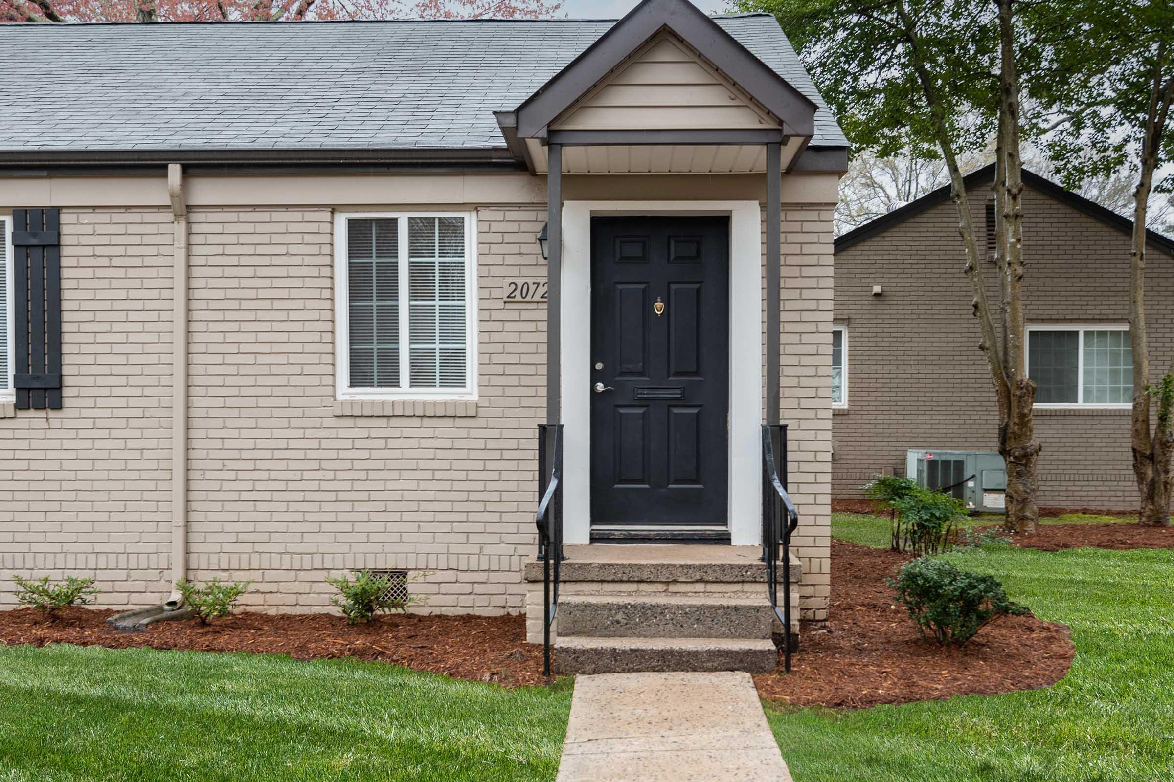 A front view of a small brick bungalow with a dark door and white window shutters. Steps lead up to the entrance, surrounded by freshly manicured grass and mulch landscaping. In the background, another building is partially visible, adding to the residential setting.