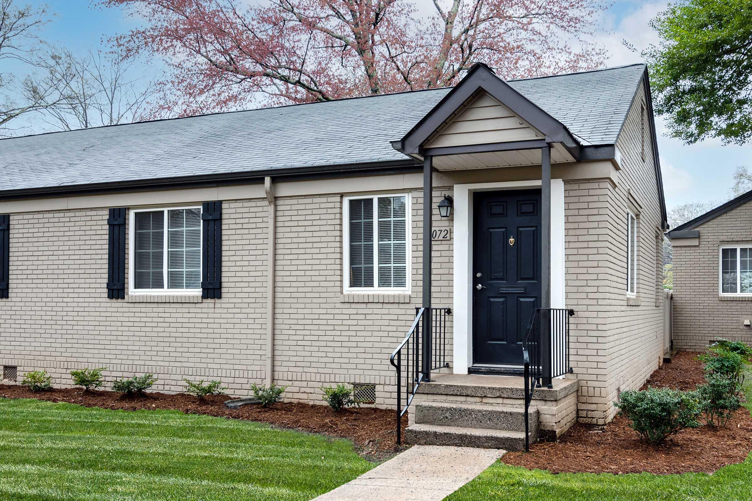 A modern single-story brick home with a black front door, white windows with black shutters, and a small set of stairs leading to the entrance. Well-manicured greenery surrounds the house, with a neat lawn and flower beds, set against a backdrop of trees.