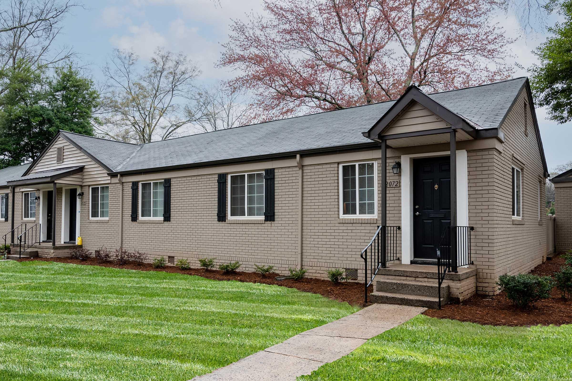A modern residential building with a brick exterior and a well-maintained lawn. It features multiple units with black shutters, a sloped roof, and a walkway leading to the front door. The landscaping includes trimmed grass and small shrubs, set against a clear sky with a few trees in the background.