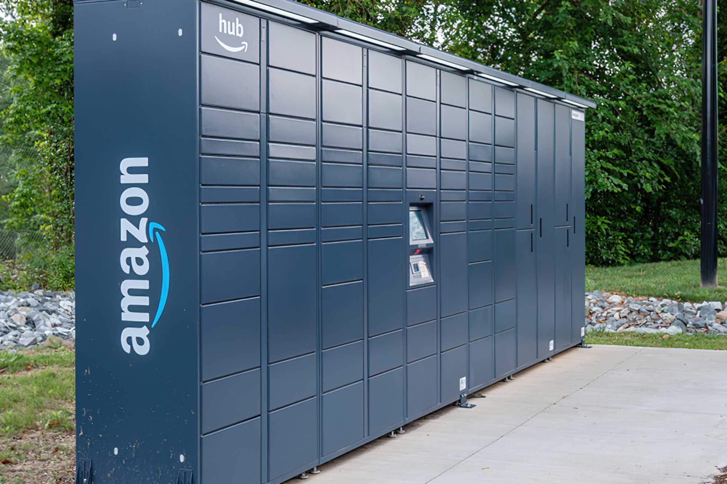 A blue Amazon Hub Locker is placed outdoors, surrounded by greenery. The locker features multiple storage compartments for package delivery, with a screen and keypad for user interaction. The Amazon logo is prominently displayed on the side of the locker.