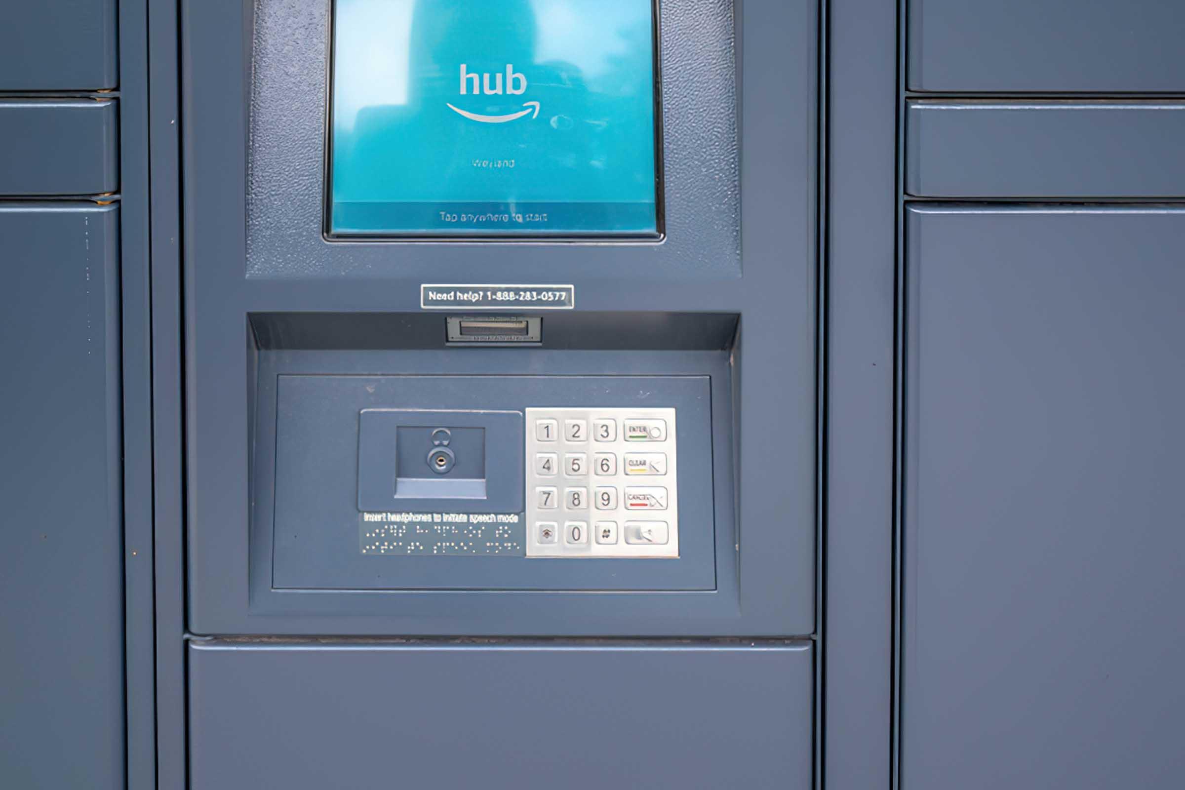 An Amazon Hub locker with a digital screen displaying the word "hub" and a keypad. Users can enter codes to retrieve packages. The locker unit is gray and features a sleek, modern design.