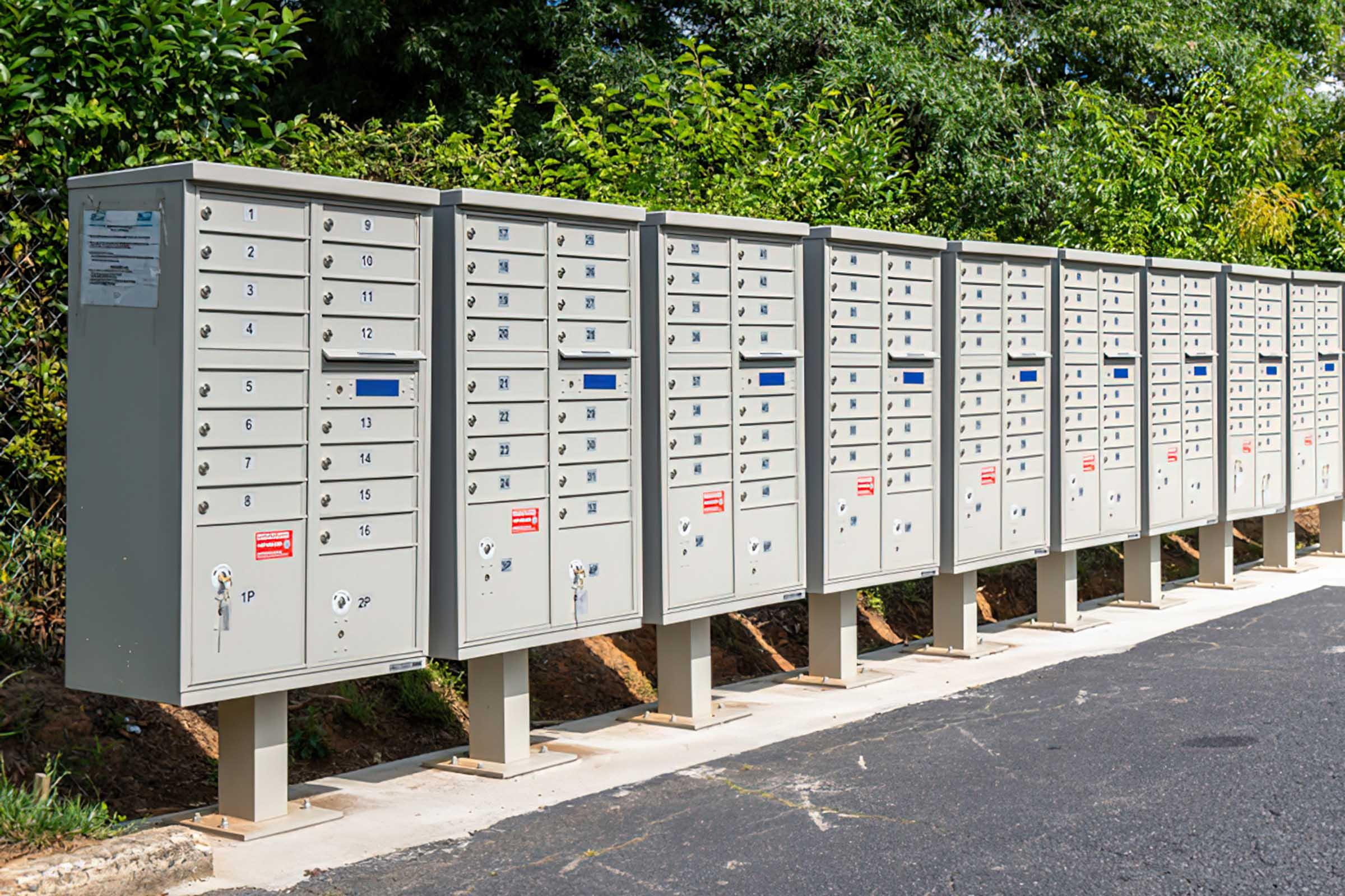 A row of multiple locked mailboxes arranged neatly along a pathway, surrounded by greenery. Each mailbox has a labeled number, and some feature blue slots for outgoing mail. The setting is well-maintained, with a paved surface adjacent to the mailboxes.