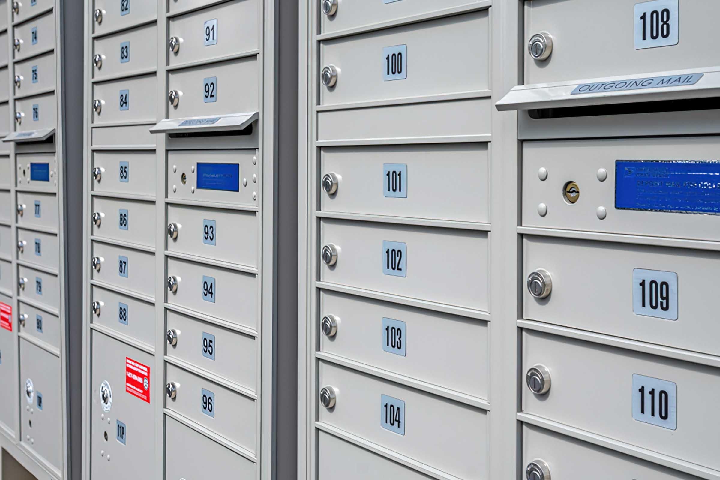 A close-up view of a series of metallic mailboxes, each labeled with numbers ranging from 91 to 108. The boxes have a sleek, modern design with individual locks and some feature blue or red labels. The photo captures multiple rows of mailboxes organized in a structured manner.