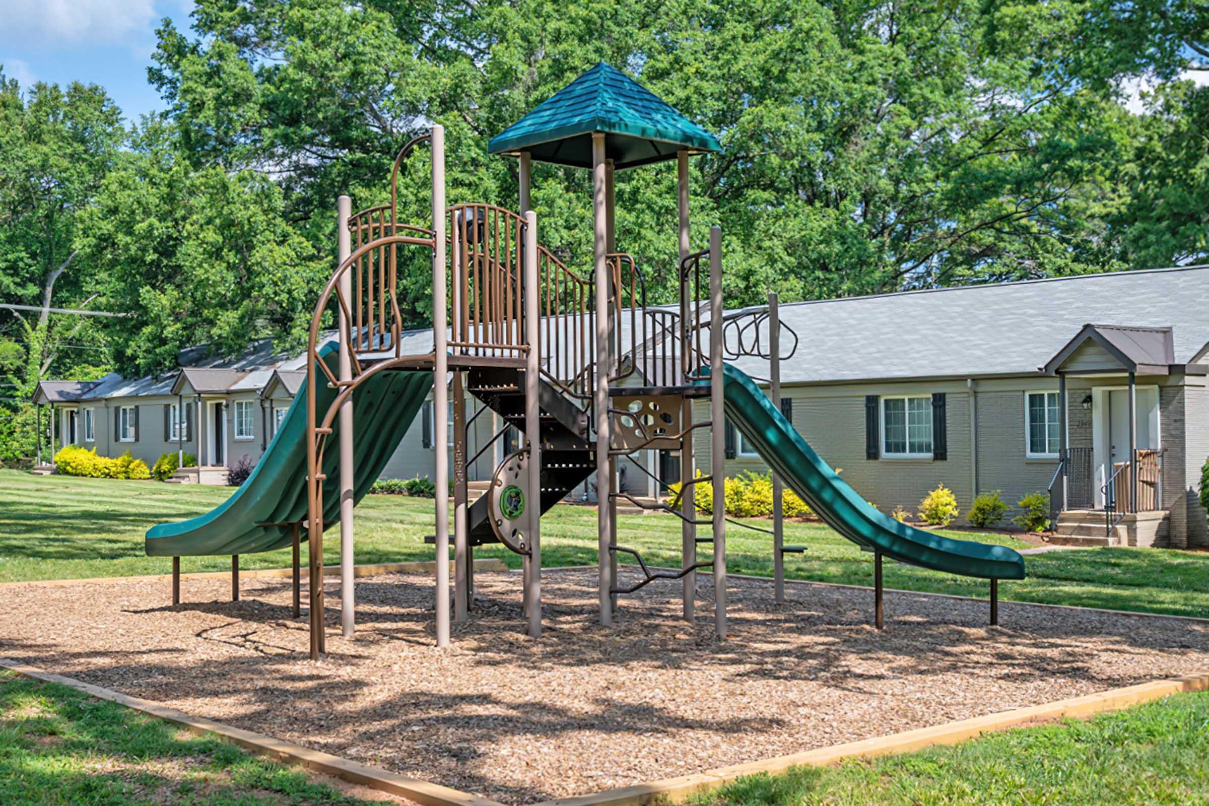 A playground structure featuring a slide, stairs, and a small tower, situated on a grassy area with mulch. In the background, there are several residential buildings with gray roofs and green landscaping, under a clear blue sky.