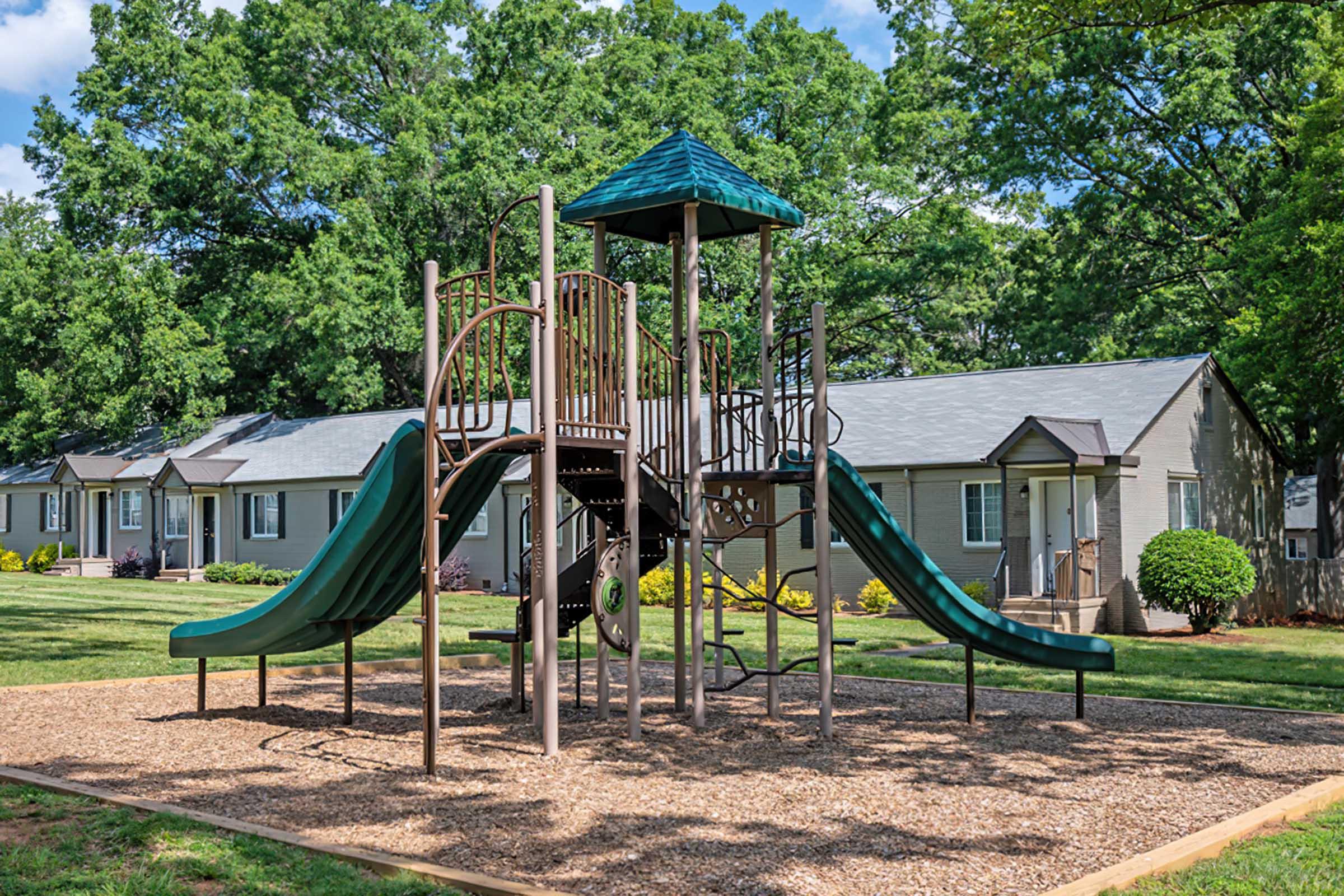 A playground featuring a multi-level play structure with two green slides, surrounded by wood chips. In the background, there are several residential buildings nestled among large trees, creating a spacious and inviting outdoor environment.