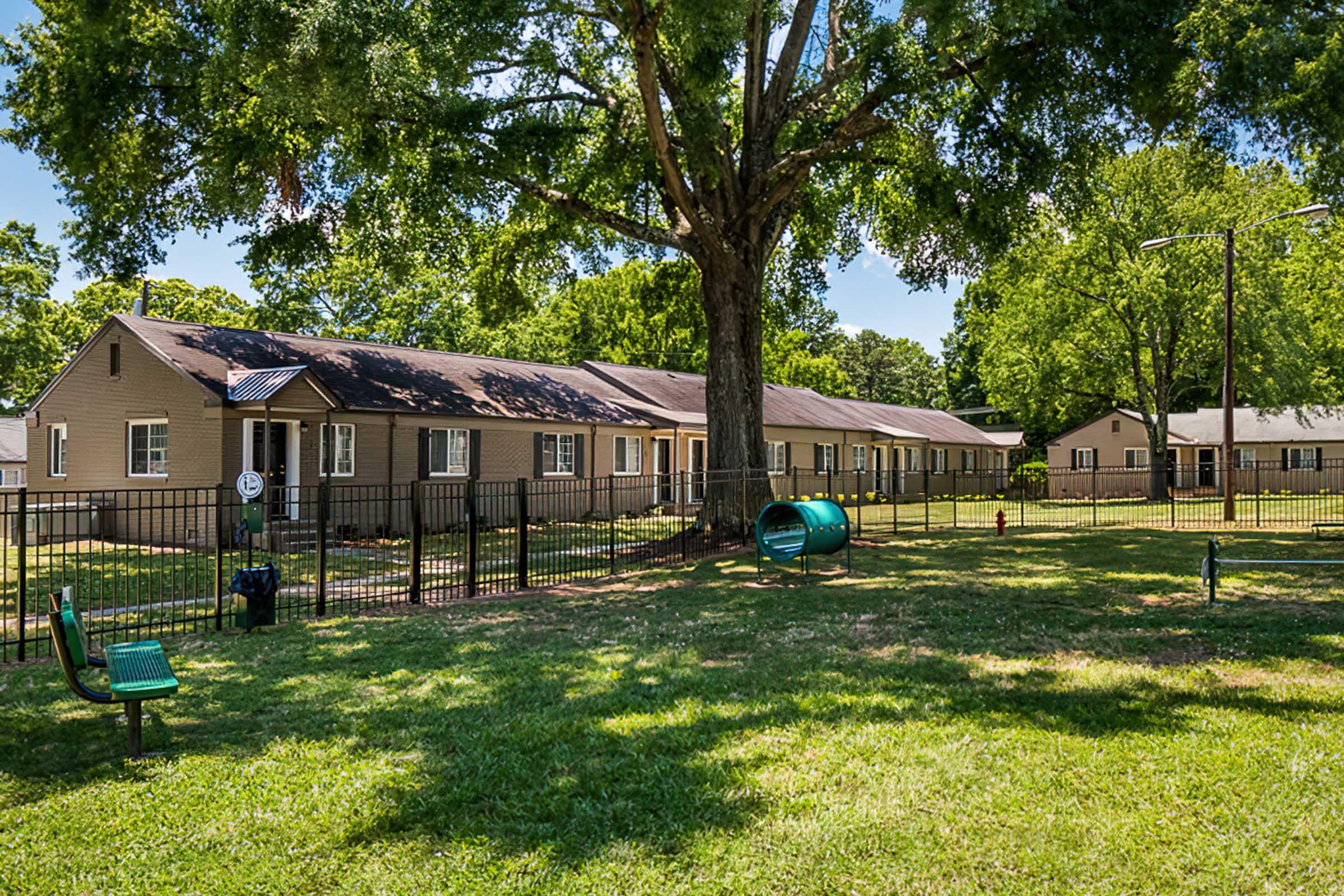 A sunny grassy area with several residential buildings arranged in a row, featuring a large tree providing shade. There are benches, a playground structure, and a fenced perimeter, creating a spacious outdoor environment for residents.