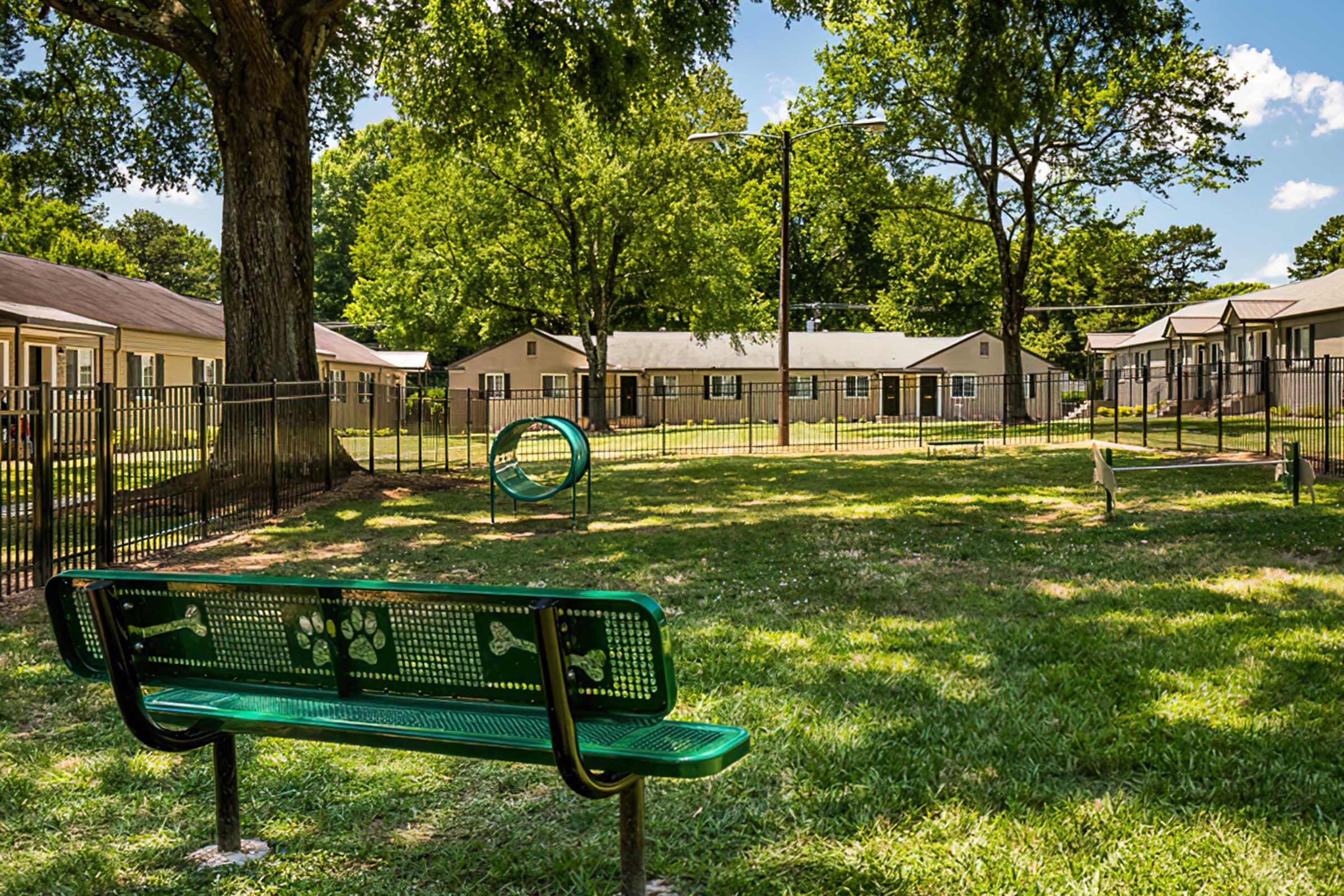 A green park bench sits on a grassy area, overlooking a fenced-in dog park with agility equipment, including a tunnel. In the background, there are several residential buildings surrounded by trees under a clear blue sky with fluffy clouds.