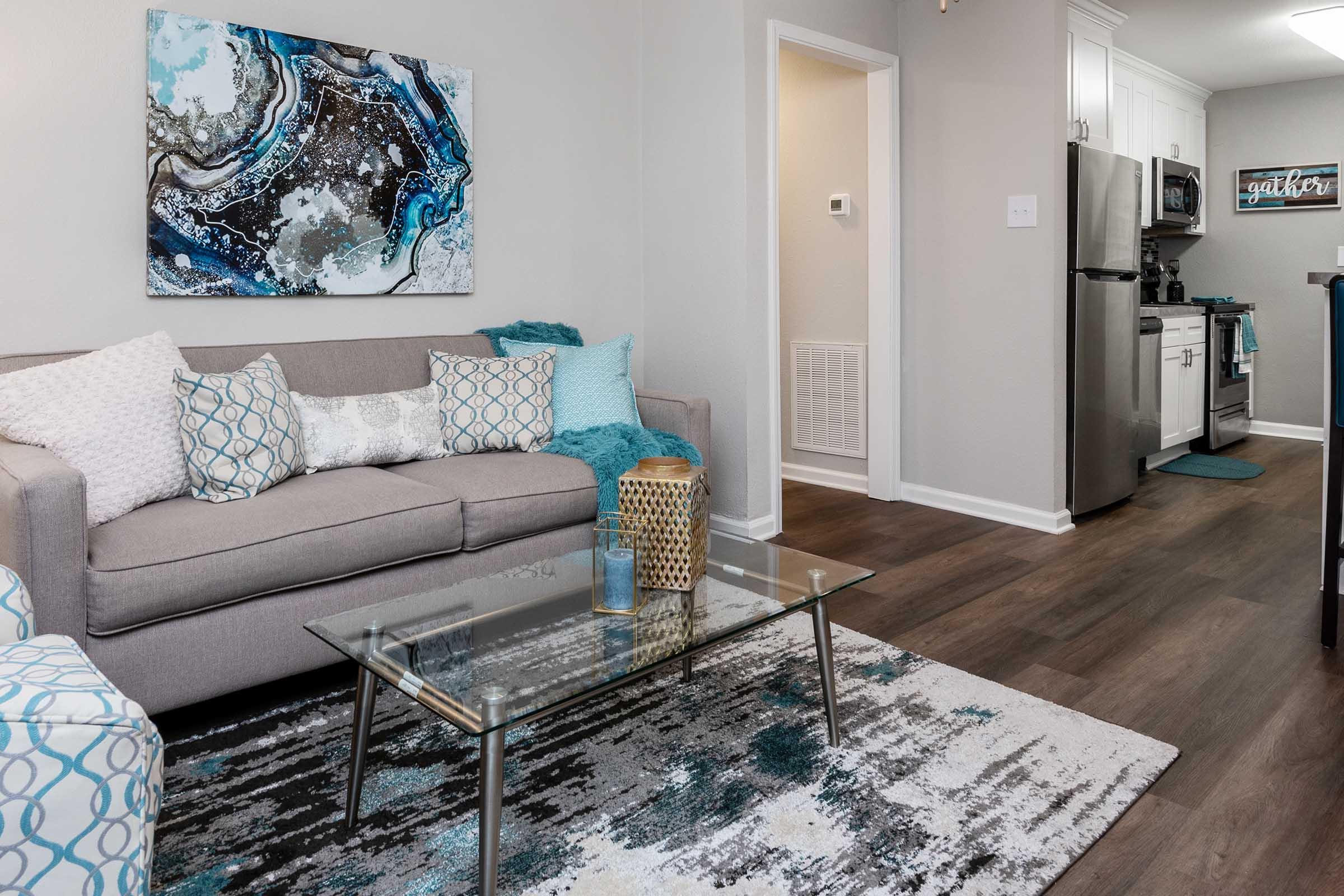 A modern living room featuring a gray sofa adorned with decorative pillows in blue and white, a glass coffee table, and a patterned area rug. The walls are painted light gray, and there’s a kitchen visible in the background with stainless steel appliances and white cabinets.