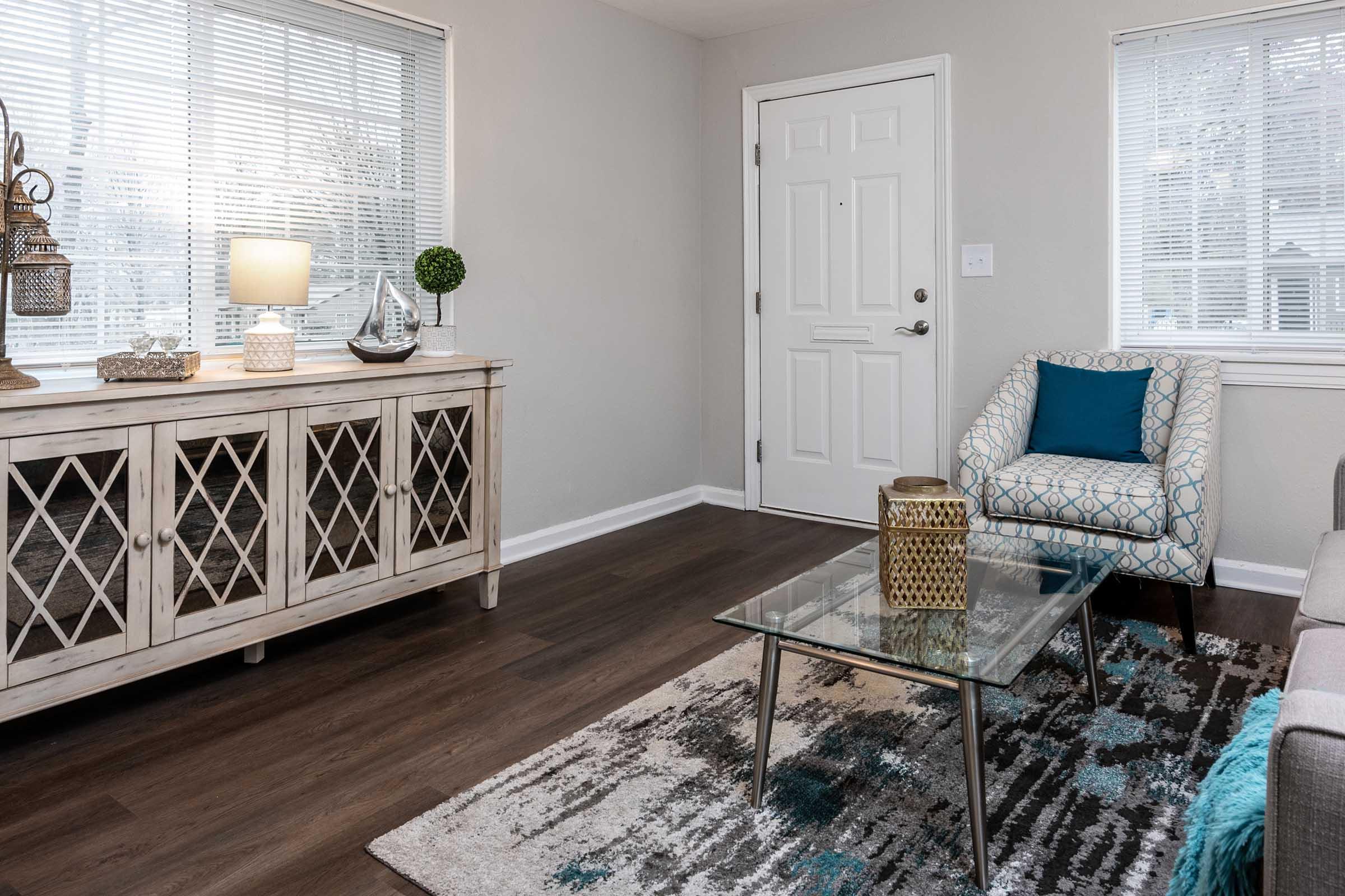 A cozy living room featuring a light-colored sideboard with a decorative lantern and small plant, a glass coffee table atop a patterned rug, and a gray sofa. There is a patterned armchair with a blue cushion near a door and two windows with blinds, allowing natural light to brighten the space.