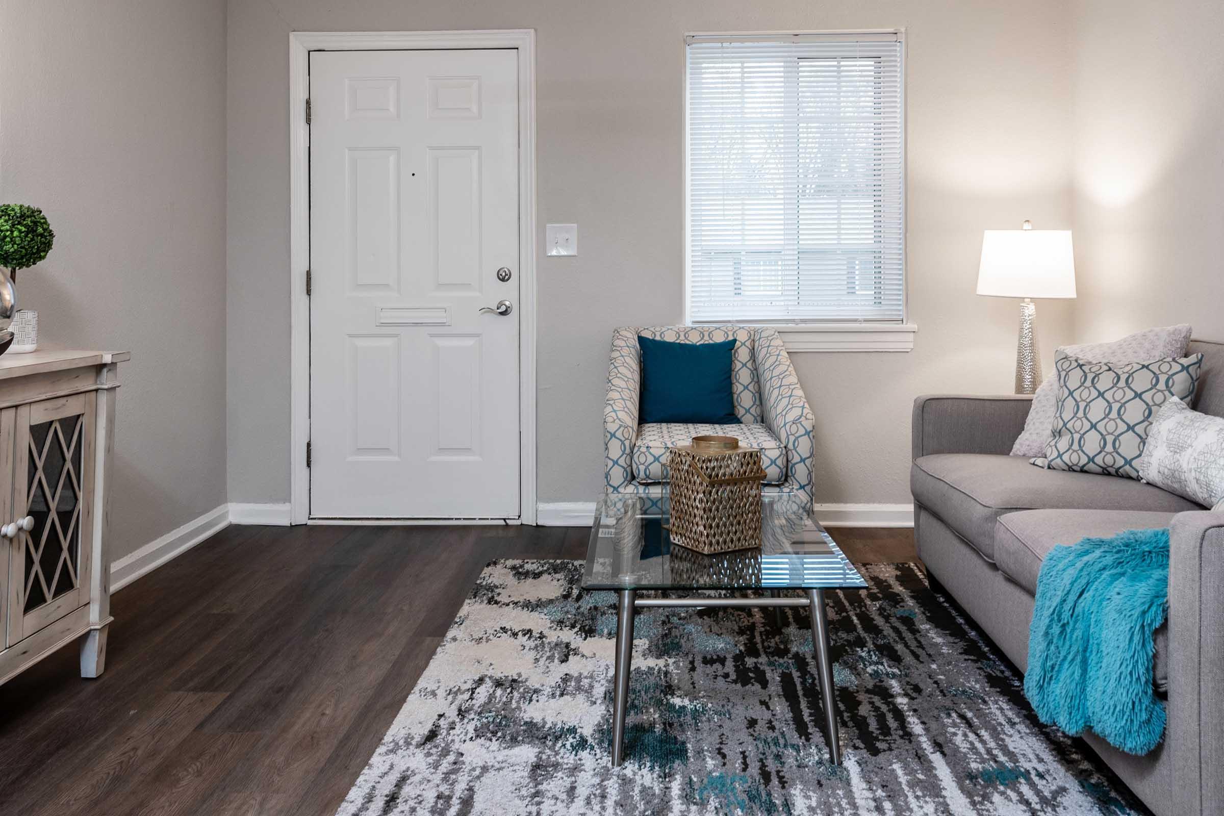 A cozy living room featuring a light gray sofa with decorative pillows, a glass coffee table with a woven basket, and an accent chair with a patterned design. There's a small table beside the chair, a lamp providing warm light, and a front door with a window above it. The floor is dark wood, and the walls are painted in neutral tones.