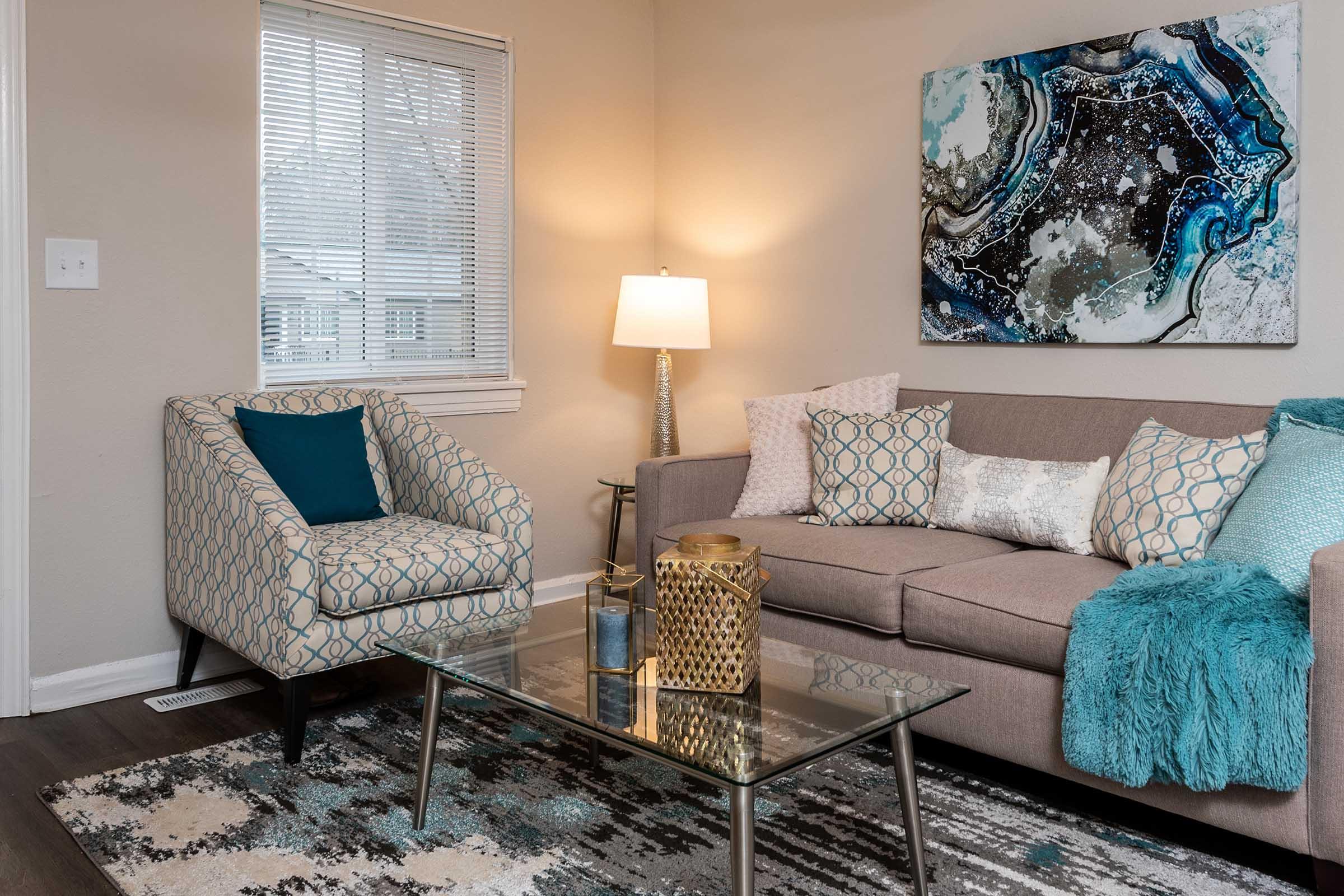 A cozy living room featuring a beige sofa adorned with decorative pillows in shades of blue, a patterned armchair, a glass coffee table with decorative items, and a warm lamp. A colorful abstract painting hangs on the wall, complemented by a soft rug underneath, creating an inviting atmosphere.